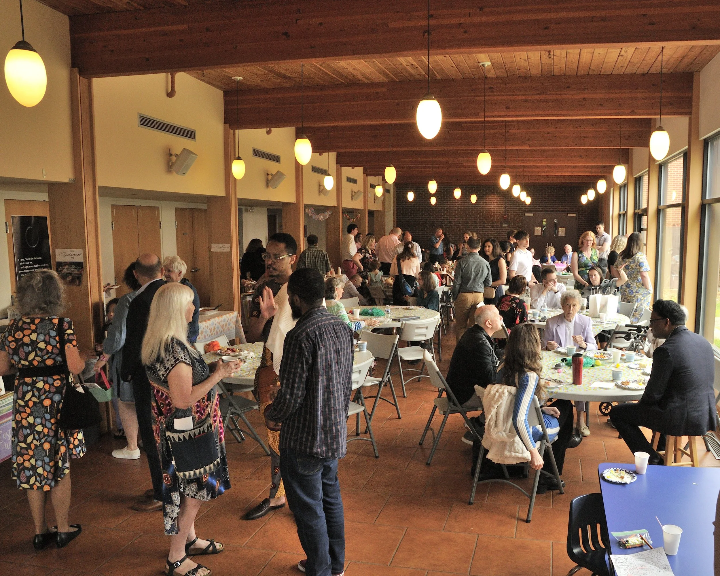 A gathering of people at an indoor event with tables, some decorated, and a wooden ceiling with hanging lights, in a room with large windows.