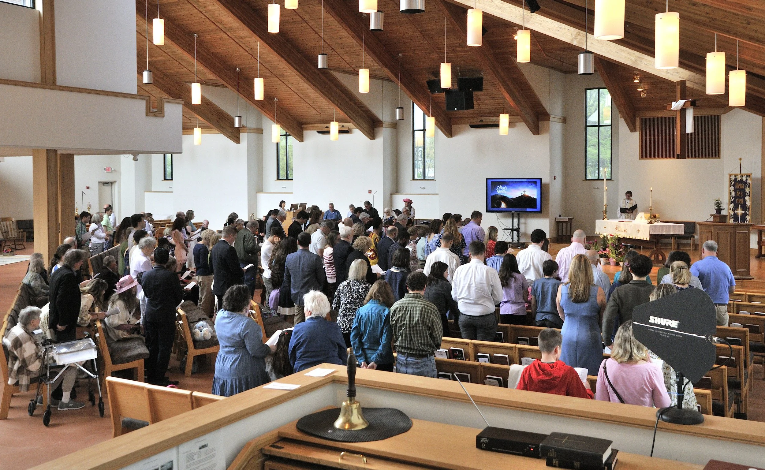 Inside a church during a service with many people standing, some sitting in pews, engaged in prayer or singing. A large screen displays religious imagery and text. The church has a wooden ceiling with hanging lights and tall windows allowing natural light, with a choir or altar area at the front.