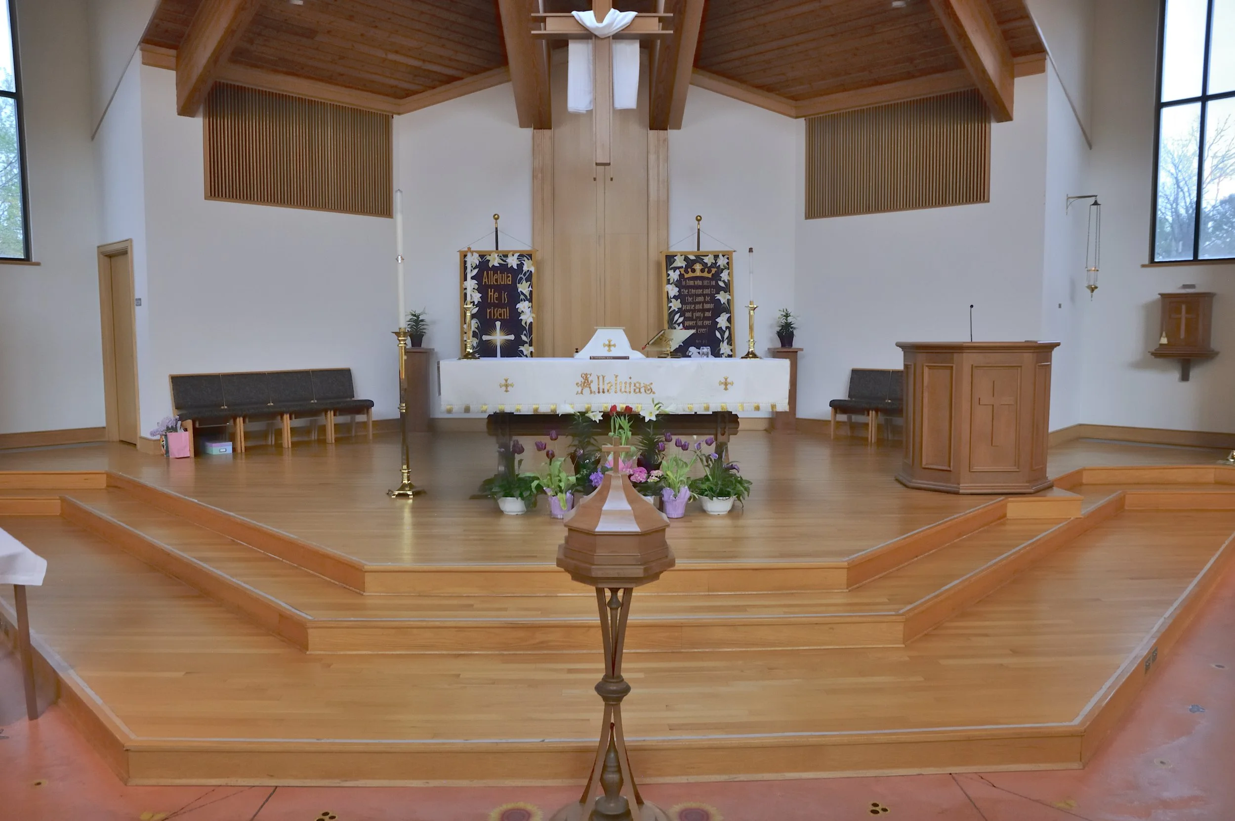 Interior of a church sanctuary with wooden flooring and ceiling, an altar decorated with flowers, candles, and religious banners reading 'Alleluia He is risen' and 'To Him who sits on the throne and the Lamb be praise and honor and glory and power forever and ever.' There are windows on both sides of the room and a wooden pulpit to the right of the altar.