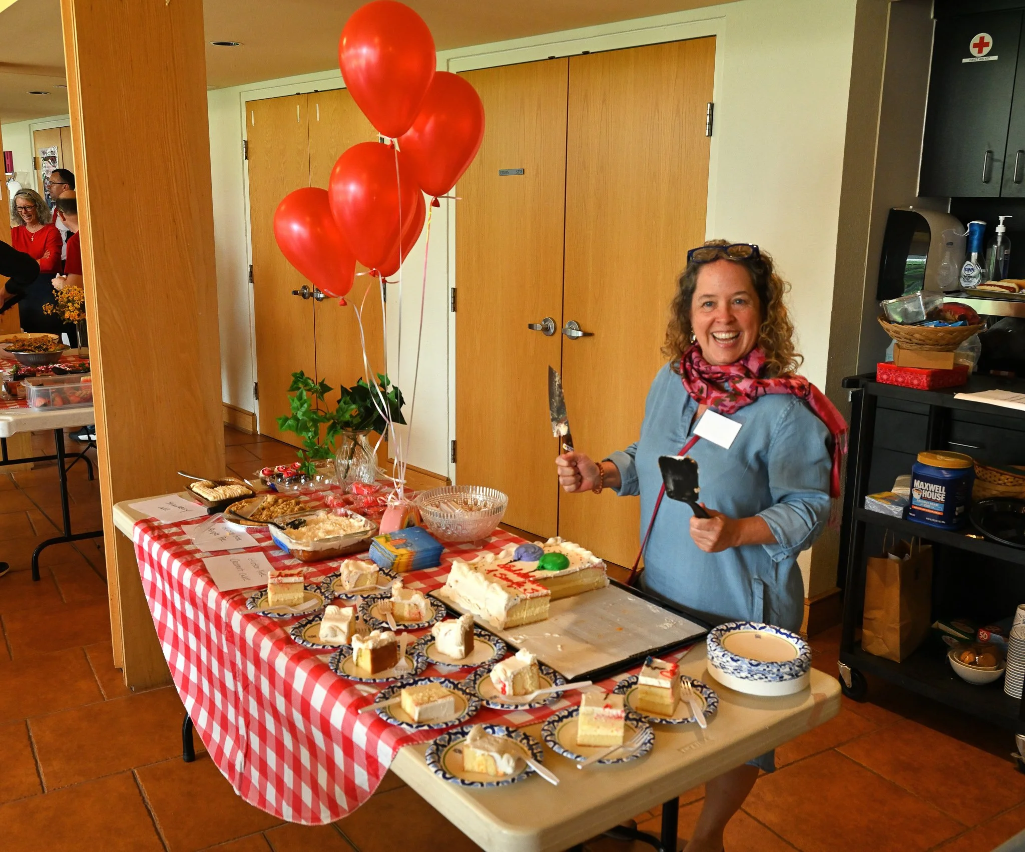 Table with assorted desserts, cakes, and snacks covered with a red and white checkered tablecloth. Red balloons are attached to a plant on the table. A woman wearing a pink scarf and glasses is smiling and holding a small knife. In the background, pe
