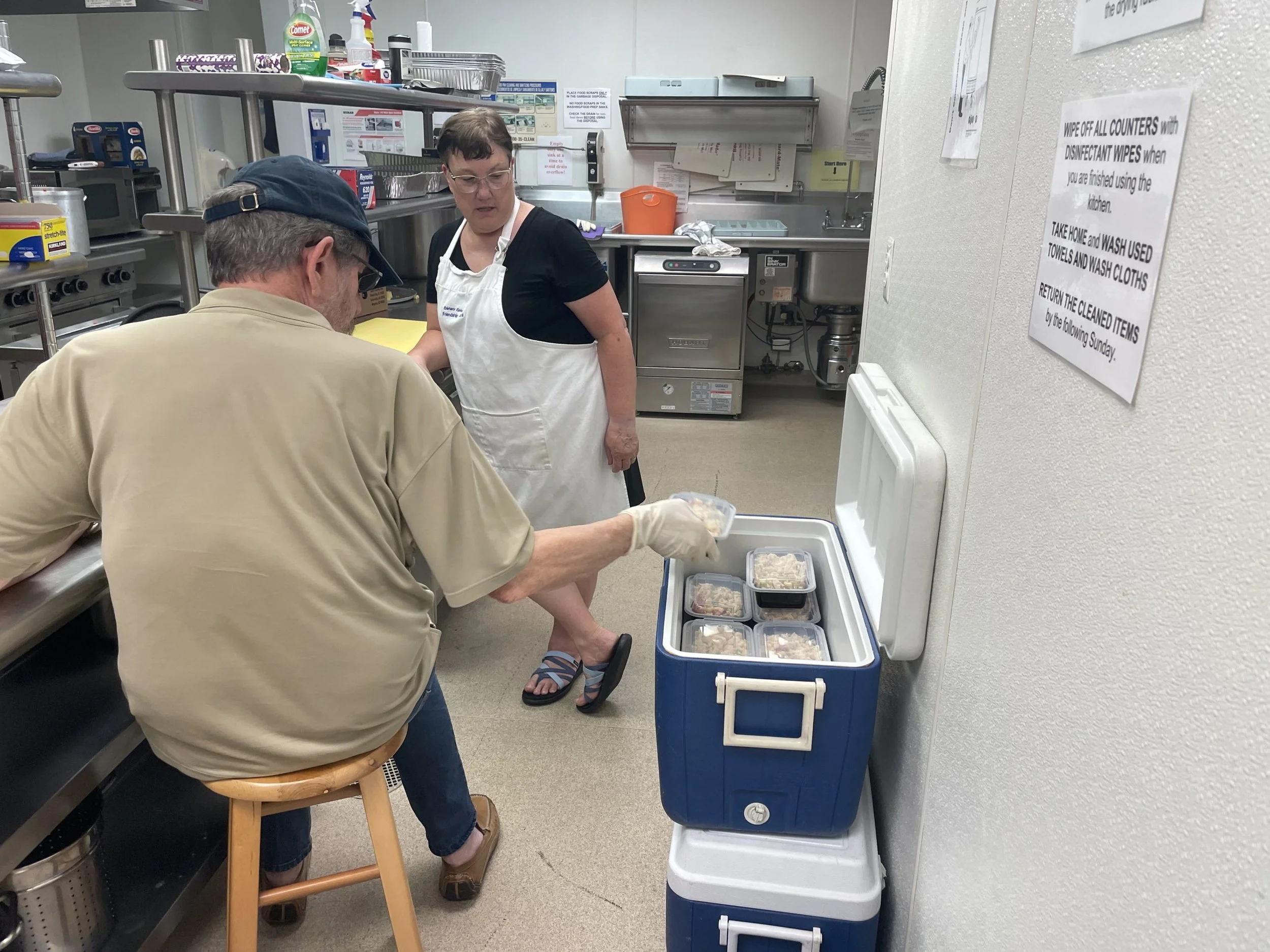 Two people are preparing food in a kitchen, with one person sitting on a stool and the other standing. There are containers with food and a cooler in the foreground, and various kitchen supplies on shelves in the background. A sink and a large white sign with instructions are also visible on the wall.