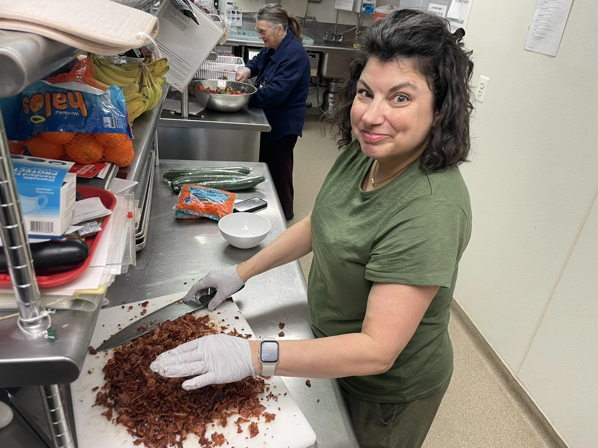 A woman preparing shredded cooked meat, possibly beef or pork, on a kitchen counter with kitchen staff in the background.