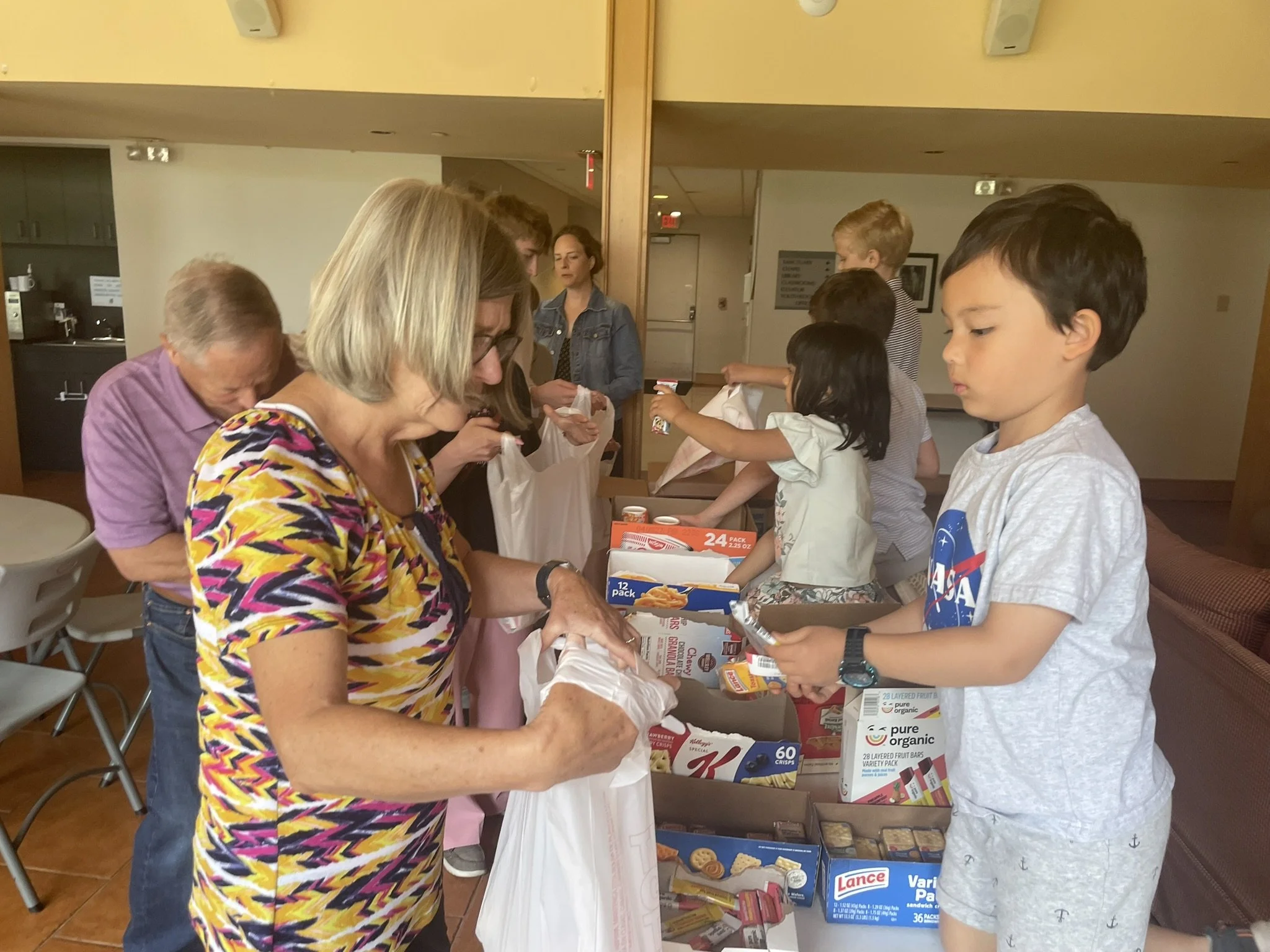 People standing in line at a table with food and snack items, including packs of snacks and boxes of crackers, inside a room with beige walls and a mirror.