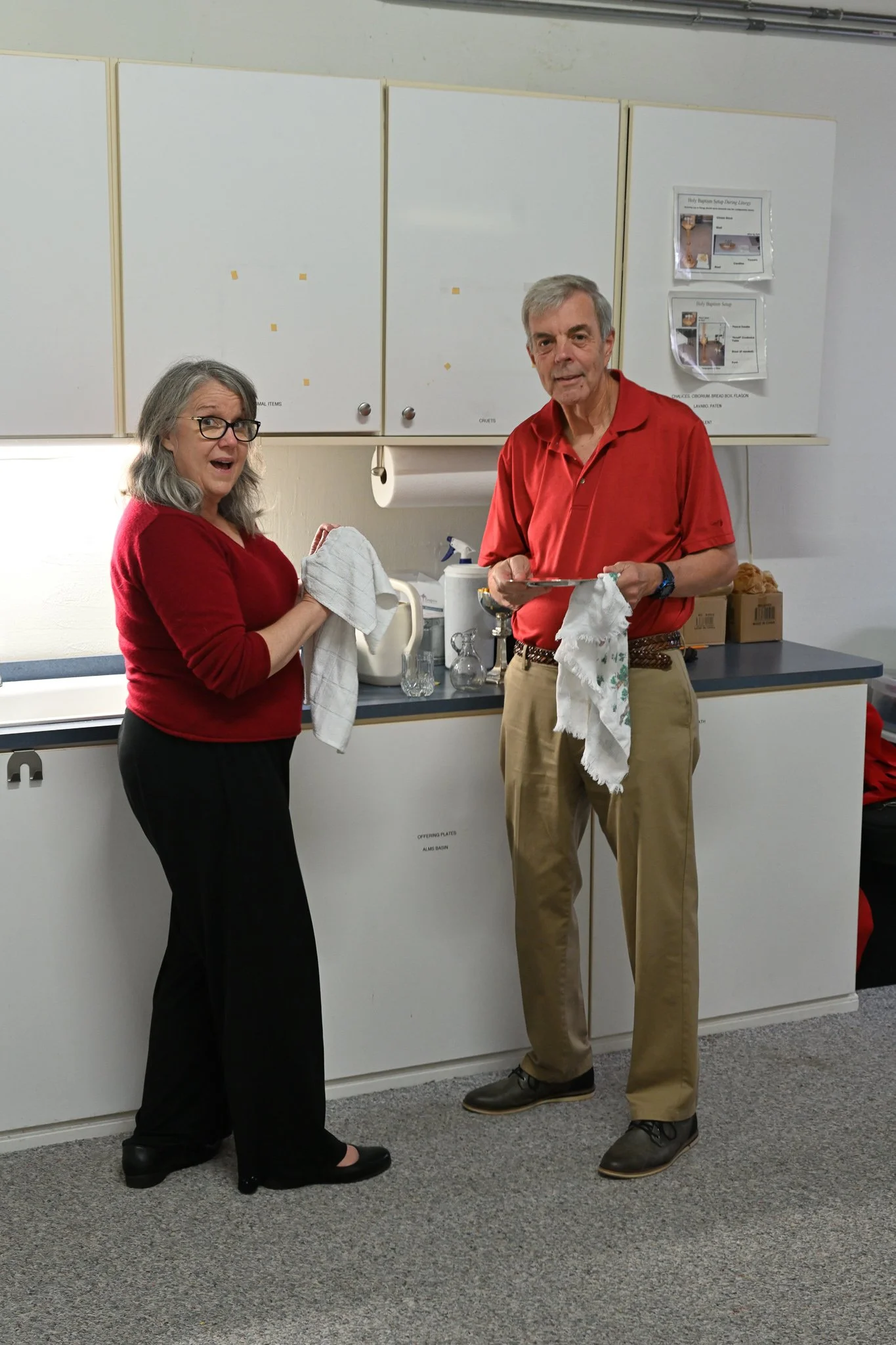  woman and a man, standing in a room with white cabinets. The woman is wearing a red sweater, black pants, and glasses, holding a white cloth. The man is wearing a red polo shirt and khaki pants, holding a dish towel. They are smiling and looking at 