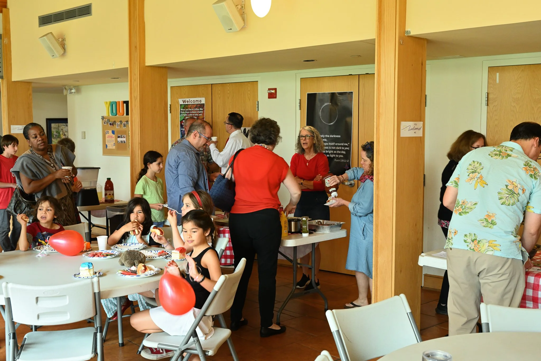 A group of children and adults gathered around a table with cupcakes and snacks, celebrating at a party in a room with wooden accents and decorated with red balloons.