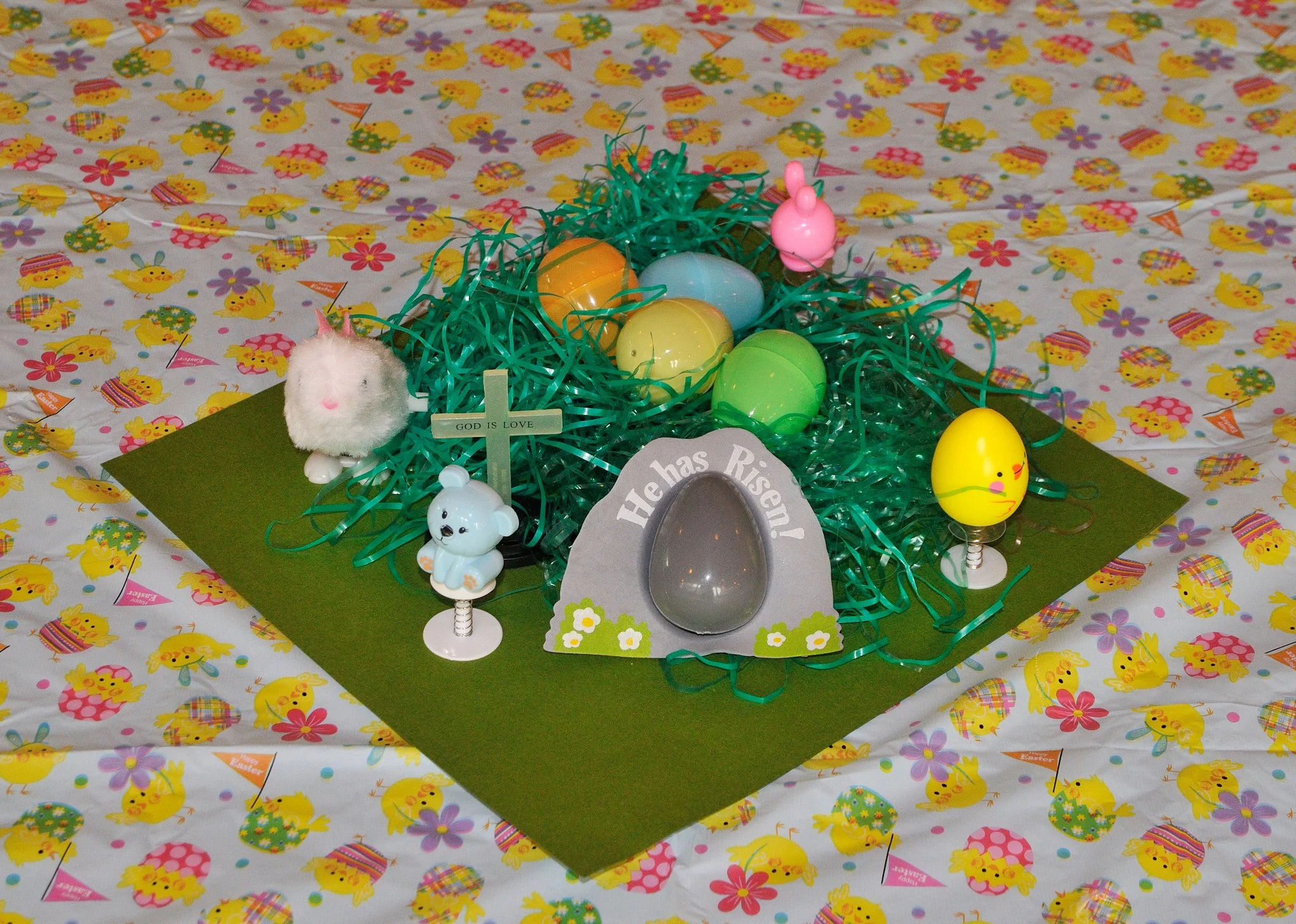 Easter-themed decorations including colorful plastic eggs, a pink bunny figurine, a small white mouse figurine, textured bunny and chick figurines, green shredded paper, and decorative grass on a table with a patterned tablecloth featuring yellow chi