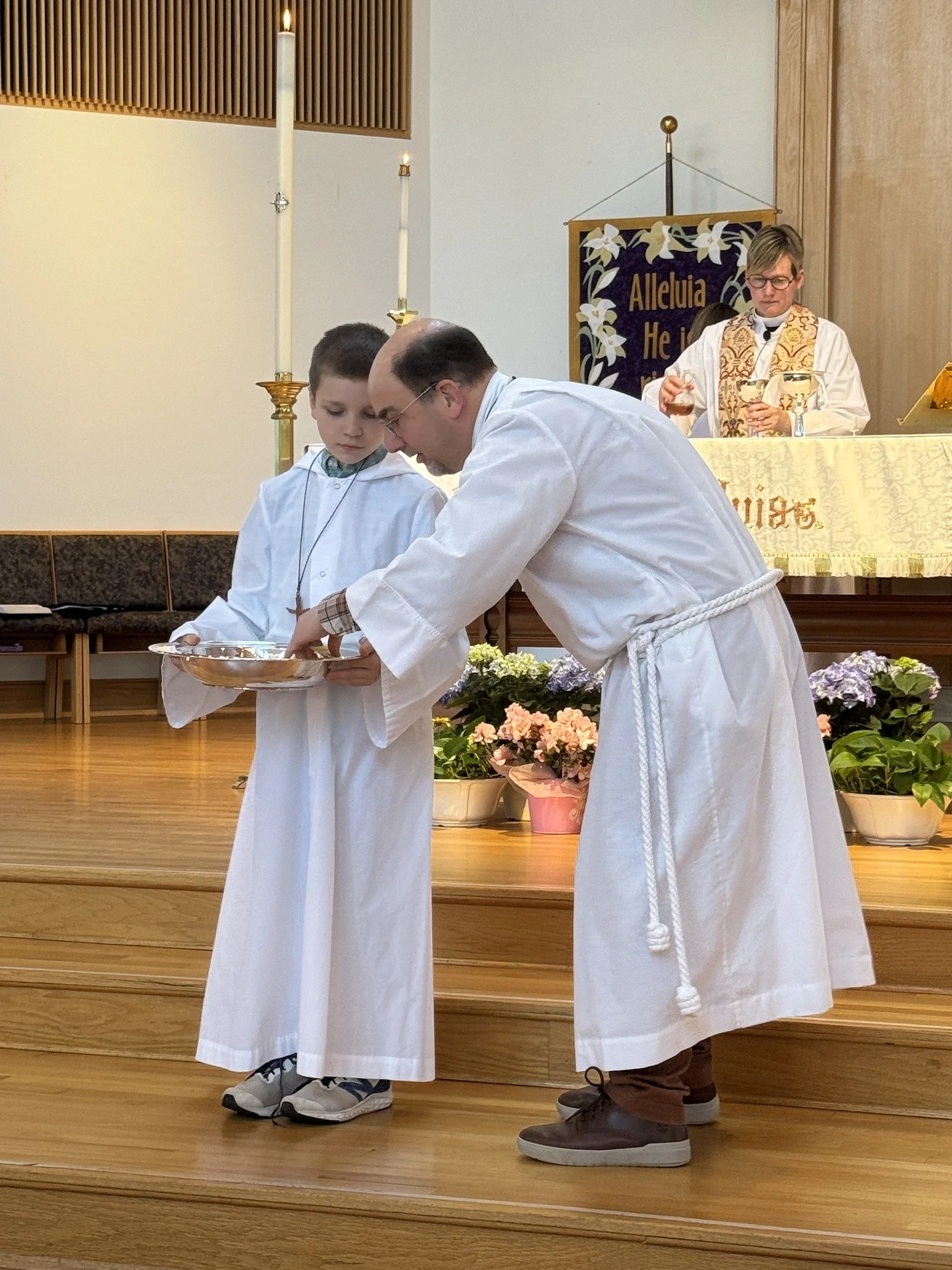 A religious ceremony in a church with a young boy and a man in white robes, both holding a silver tray, with a priest or minister in the background at an altar decorated with flowers and a banner reading 'Alleluia He is.'