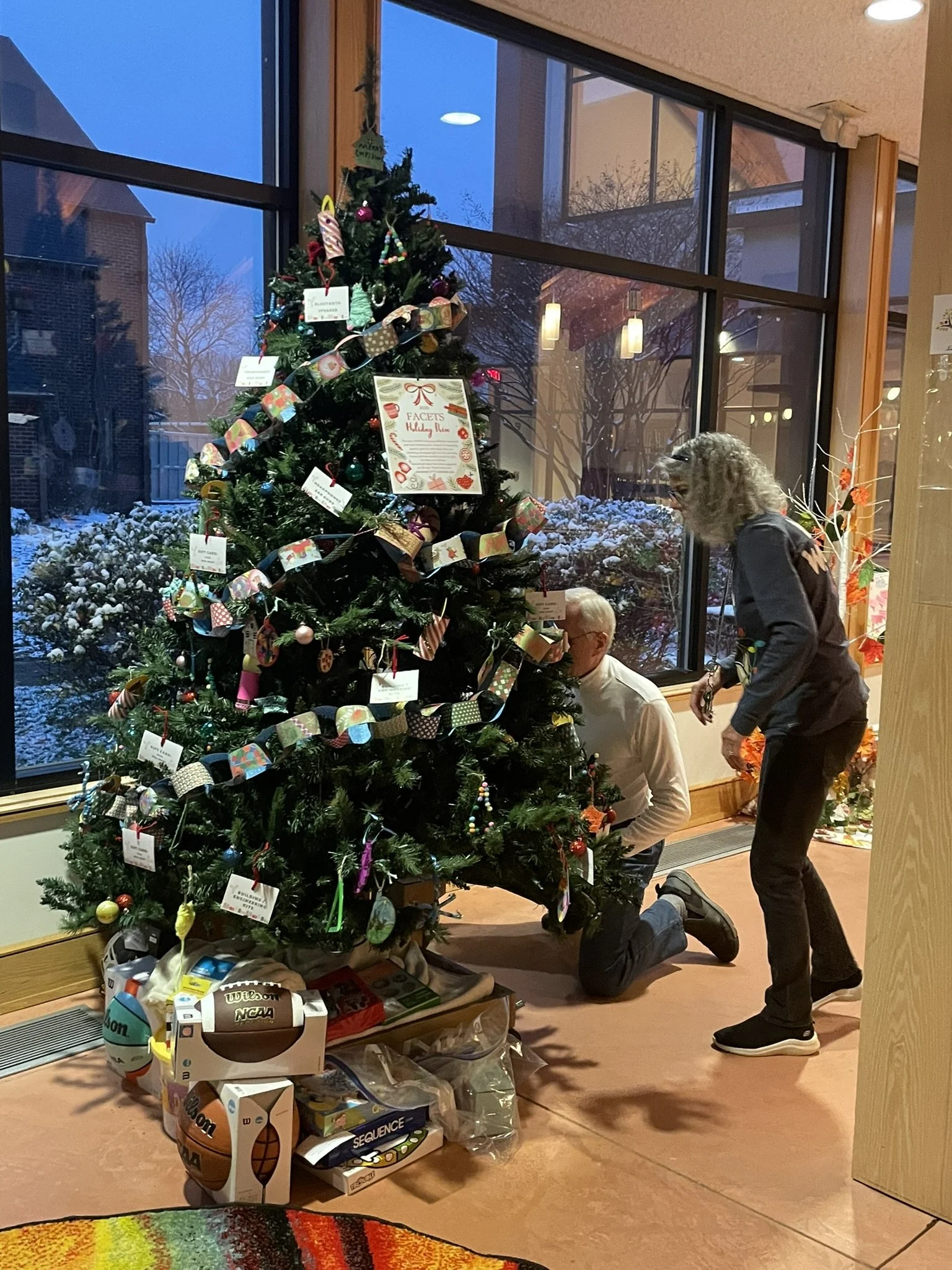 A decorated Christmas tree with ornaments and ribbons inside a building near large windows. A woman is kneeling and a man is standing nearby, both in front of the tree. Outside, snow covers the ground and trees. Items like sports balls, games, and gifts are on the floor in front of the tree.
