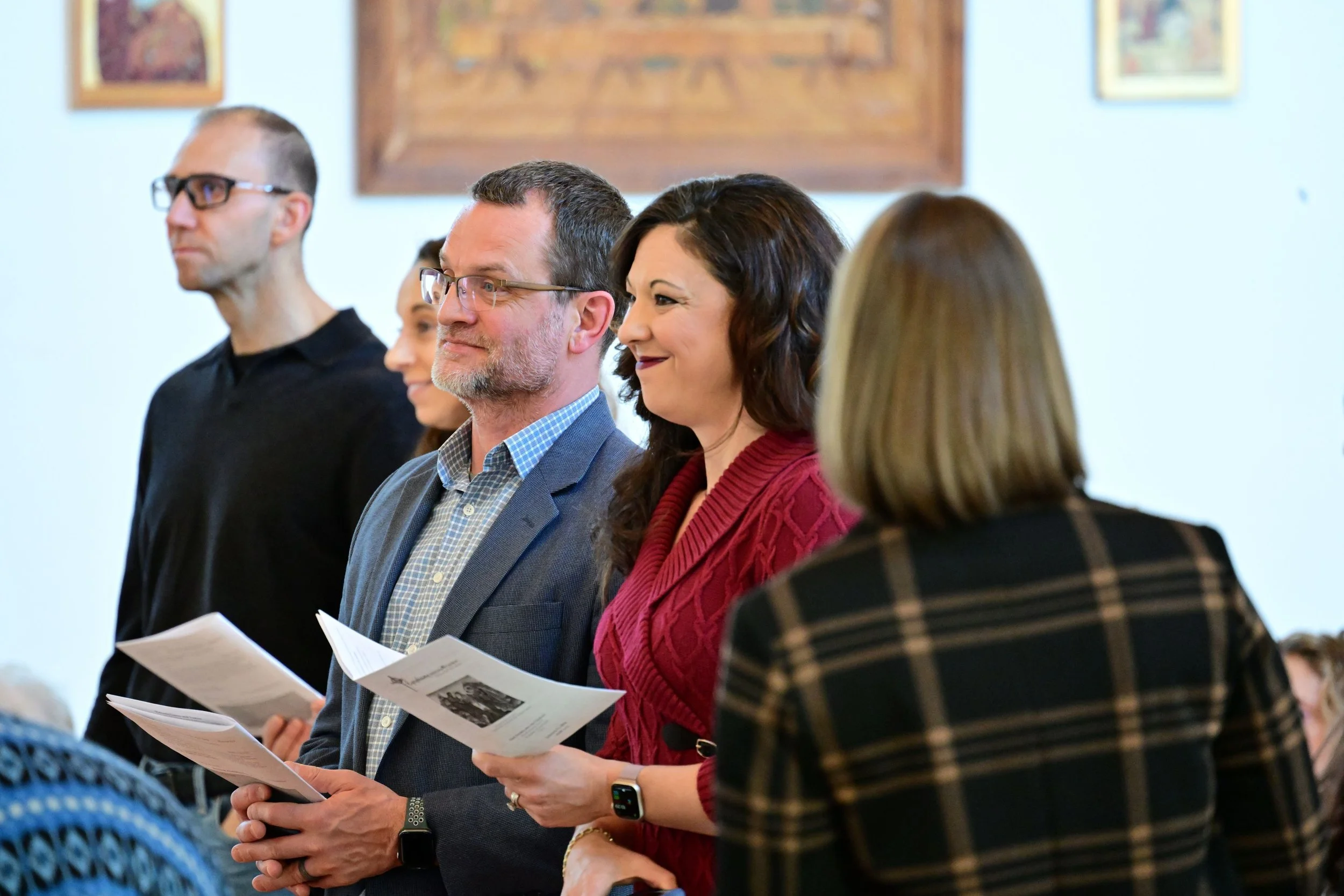 A group of people standing in a row holding papers, likely participating in a formal event or ceremony, indoors with framed artwork on the wall behind them.