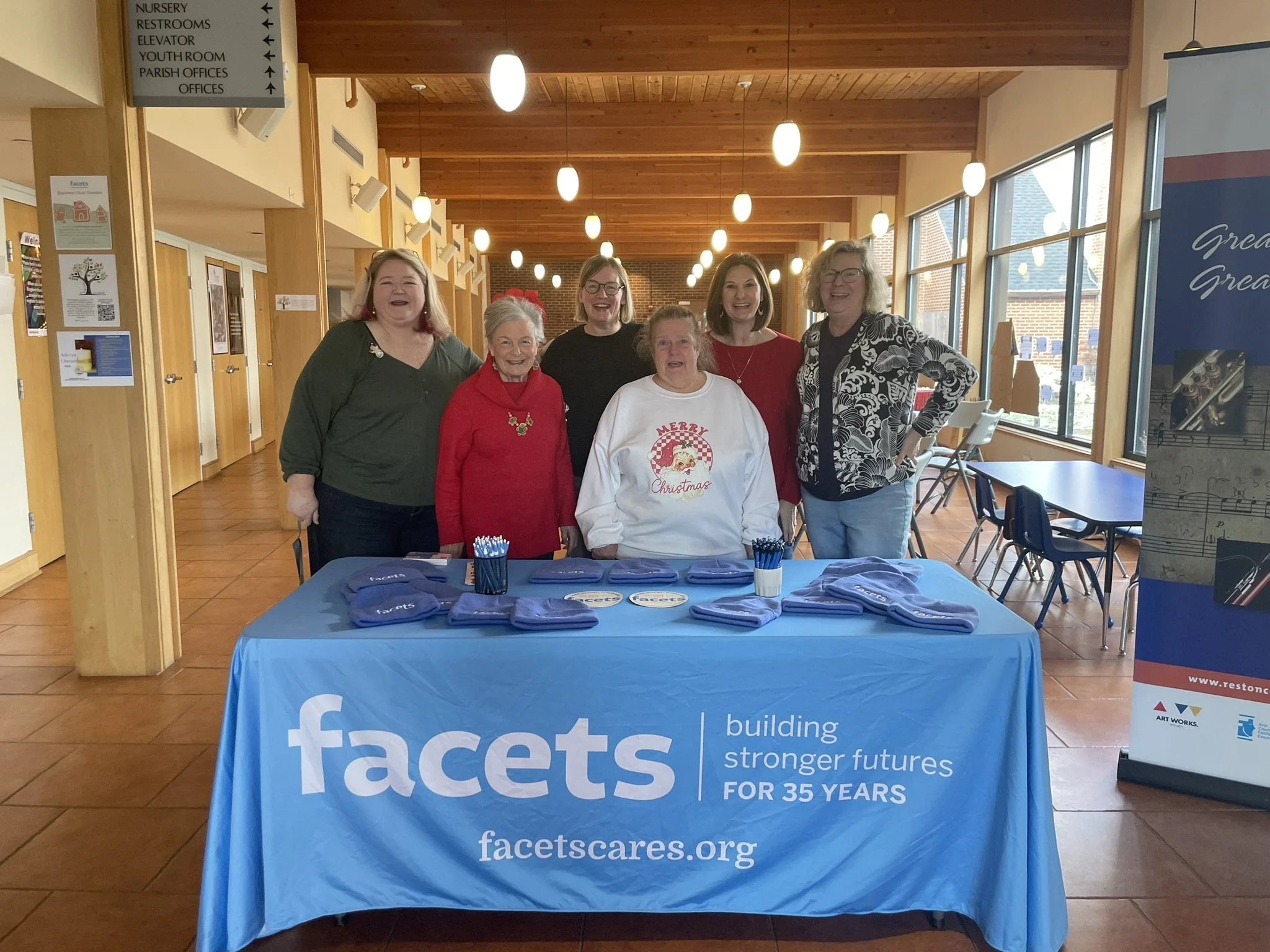 Group of six women standing behind a table with a blue tablecloth that has 'facets' printed on it, inside a building with wooden ceilings and large windows, promoting Facets organization.