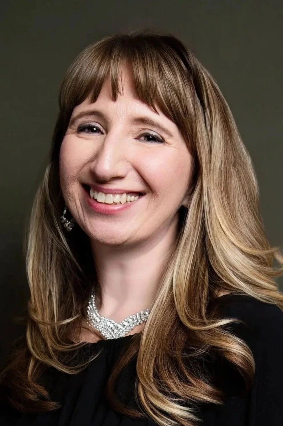 A woman with long, wavy, light brown hair, smiling, wearing a black top, pearl necklace, and earrings against a dark background.
