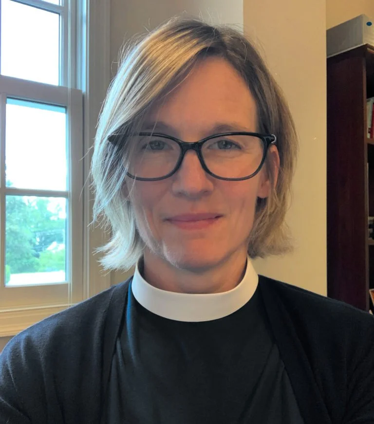 A woman wearing glasses and a clerical collar, smiling in a room with a window showing green trees outside.