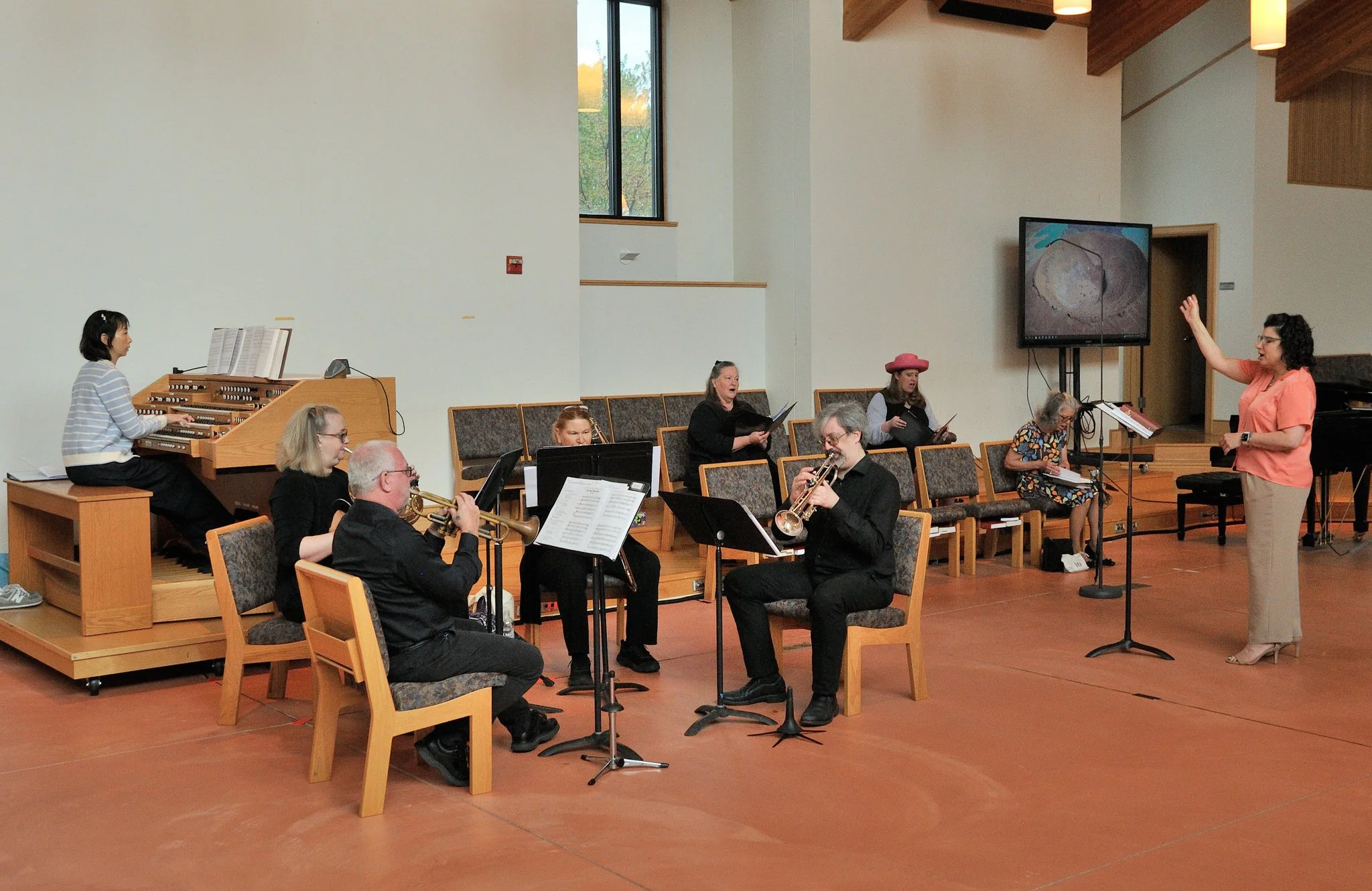 A choir and musicians rehearsing in a church or auditorium, with a woman conducting and others playing instruments or singing, some standing and some seated, with a large screen displaying an image behind them.