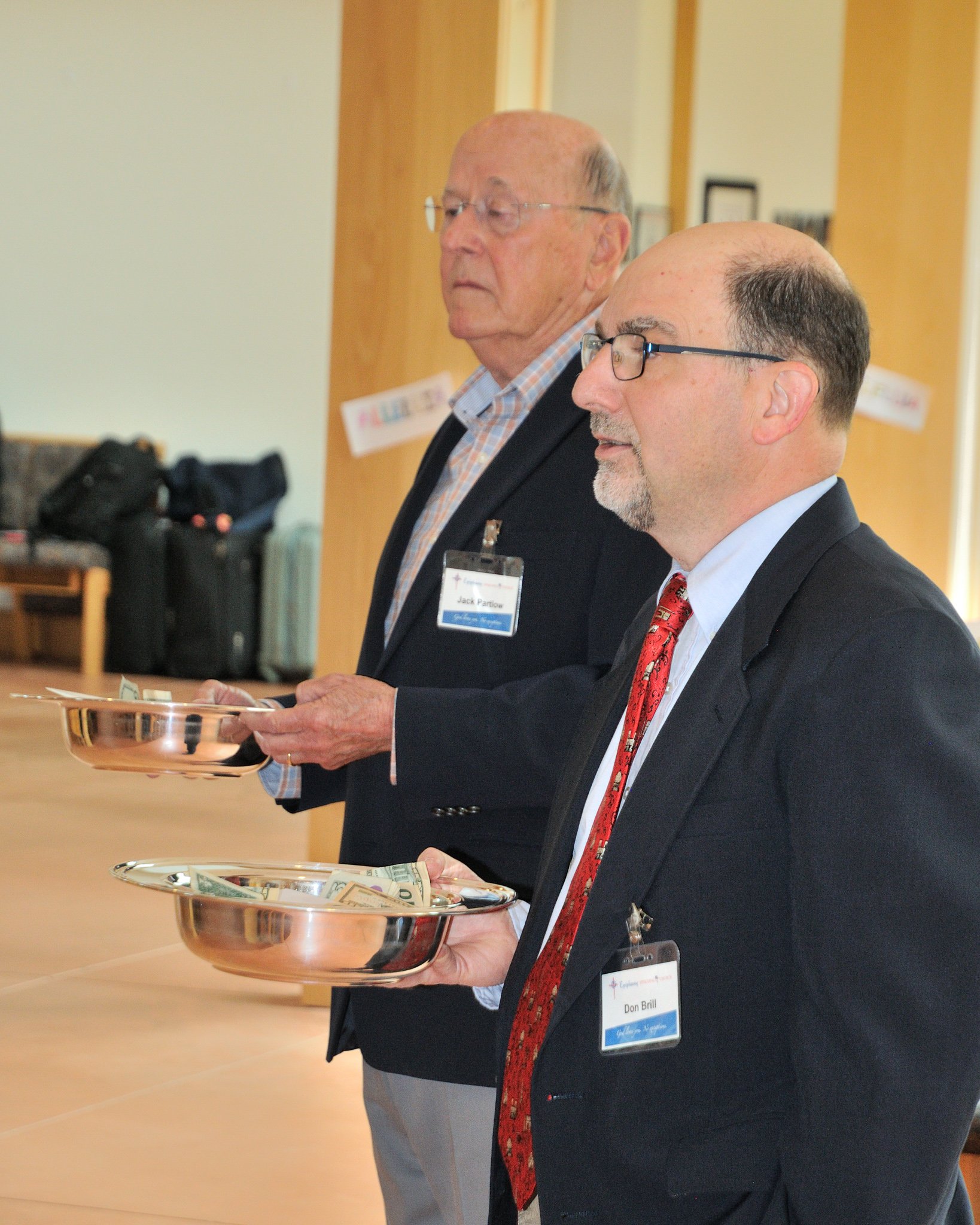 Two men in suits holding metal bowls with bills. The man in the foreground has a wig and glasses, wearing a red tie with a pattern, and a name tag reading Don Brill. The man in the background has a wig and glasses, wearing a checked shirt under an op