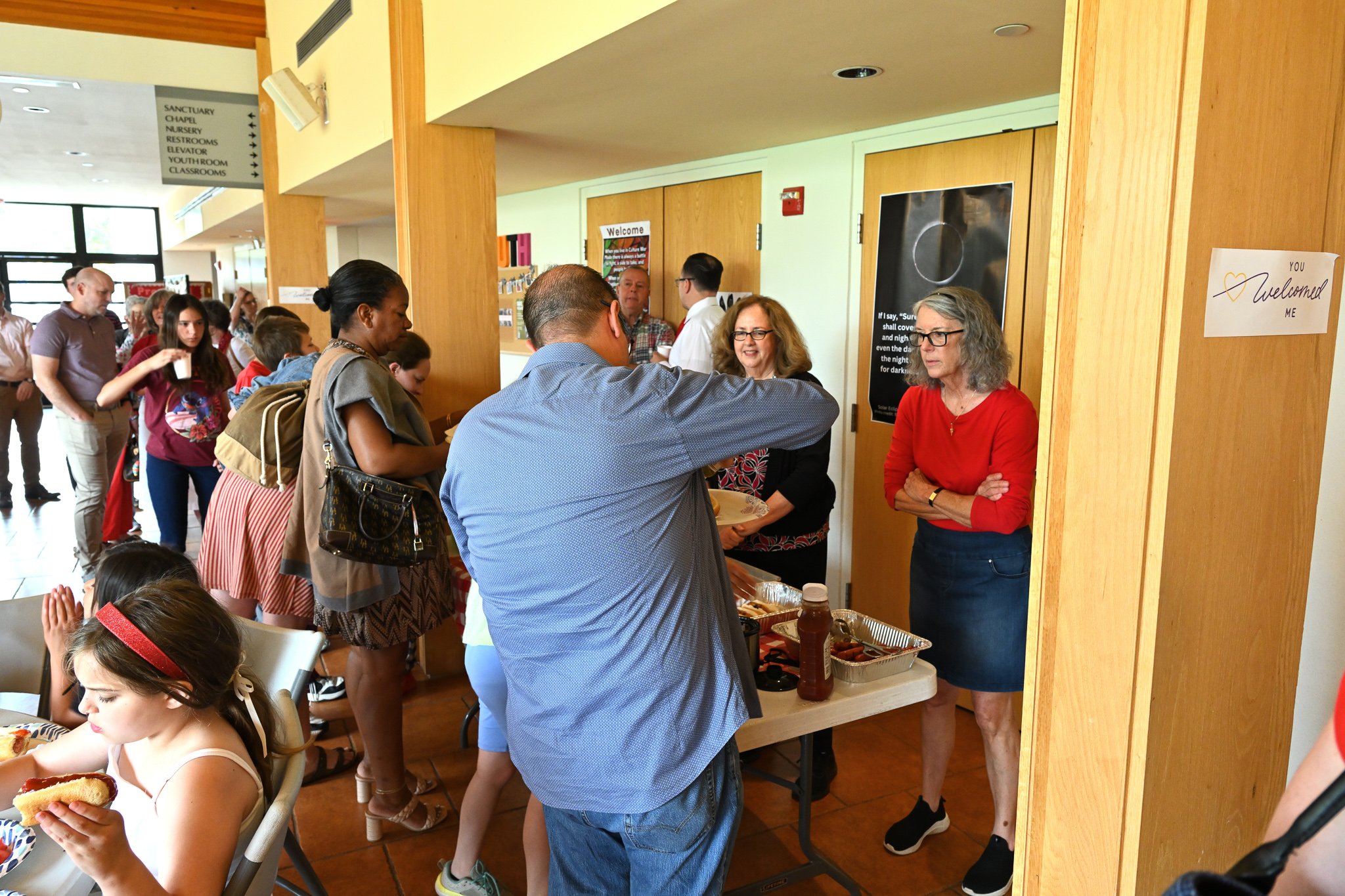 People in line at a food or service table inside a building, with signs, walls, and windows in the background.