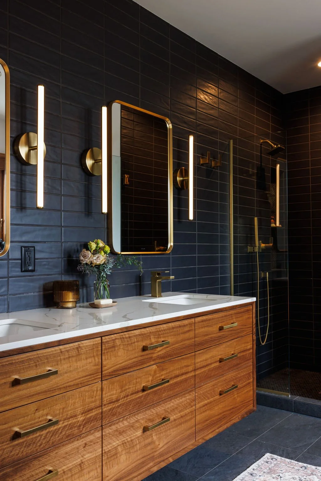 Modern bathroom with dark tiled walls, wooden vanity with gold handles, white marble countertop, and gold fixtures. It features two lit vertical mirrors, a vase with flowers, a glass candle holder, and a glass-enclosed shower with gold accents in the