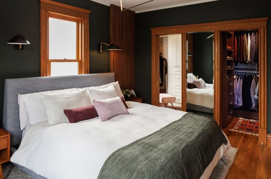 A bedroom with dark green walls, a large bed with white bedding, pillows, and a gray headboard, wooden window frame, and closet with mirrored doors showing clothes and drawers.