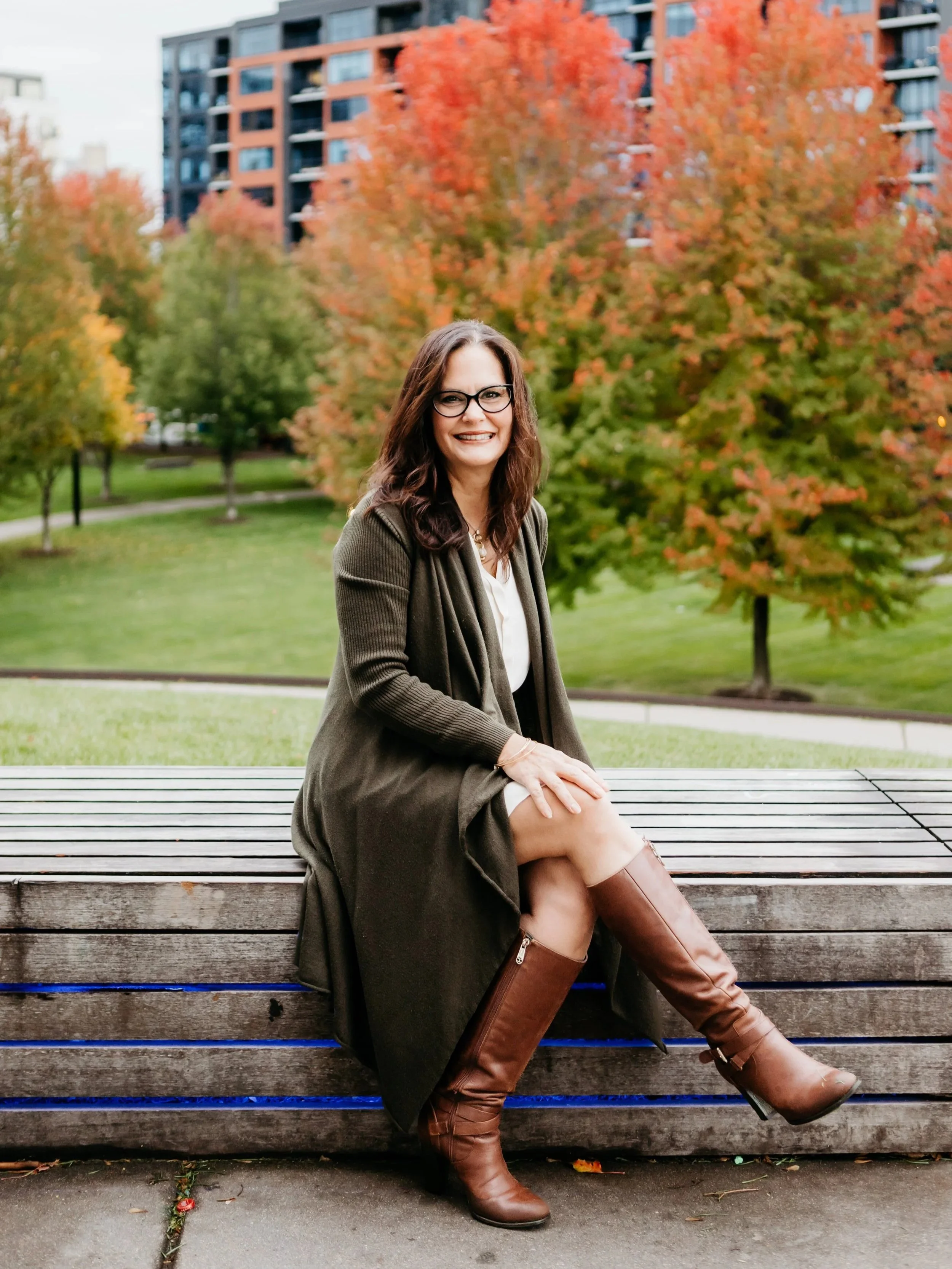 A woman with brown hair, glasses, and a dark green coat sitting on a park bench during fall with colorful orange and green trees and a multi-story building in the background.