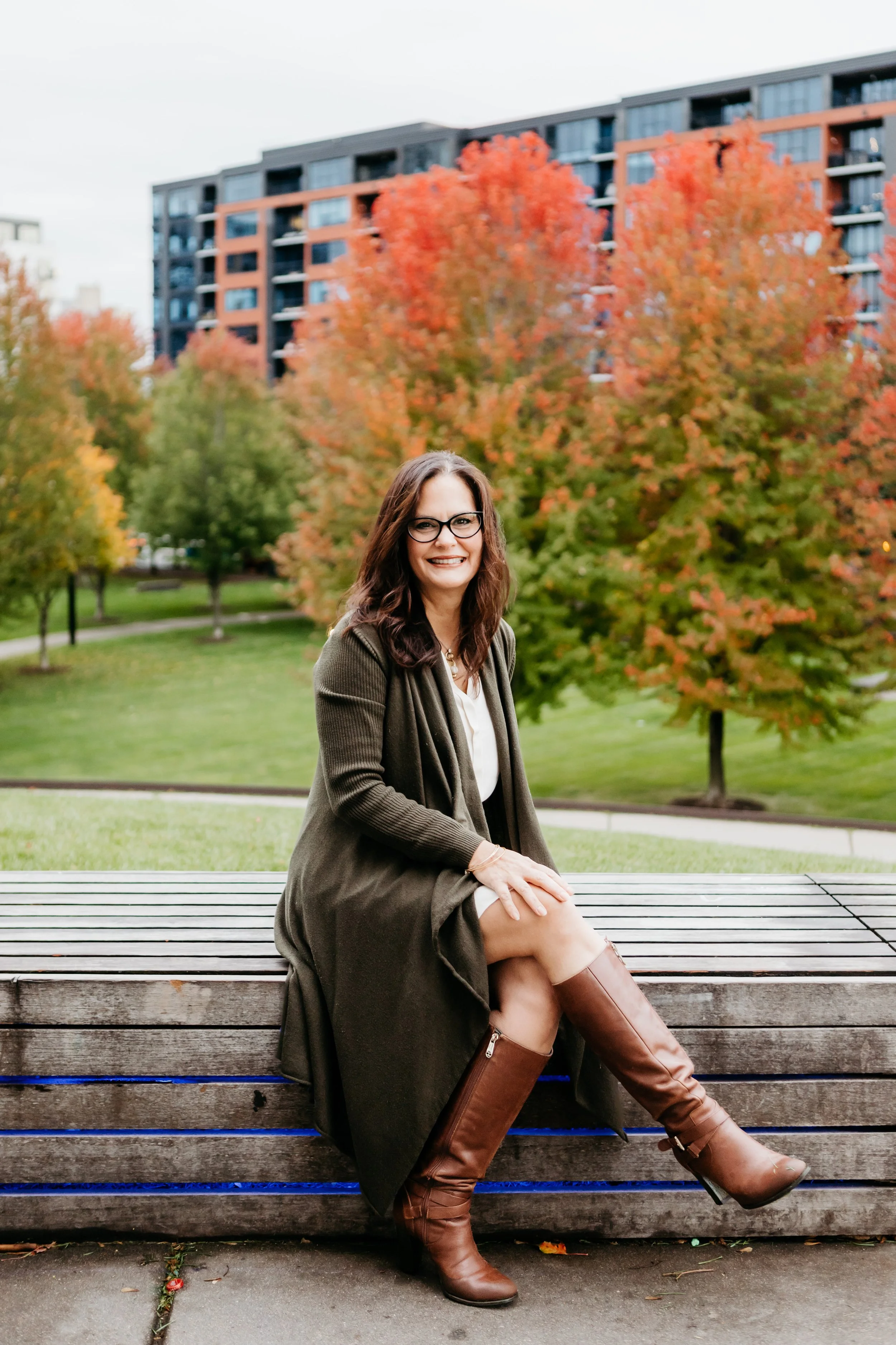 A woman sitting on a wooden bench outdoors, with colorful autumn trees and a modern apartment building in the background.