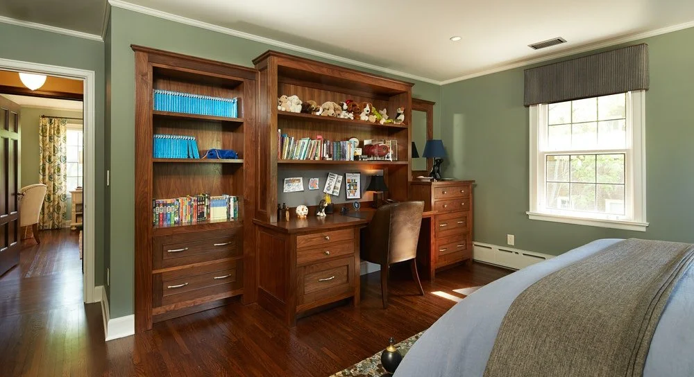 A bedroom with green walls, a wooden desk, and a bookcase filled with books and stuffed animals. There is a brown chair at the desk, a table lamp, and a window with a gray valance. Part of a bed with light-colored bedding is visible in the foreground