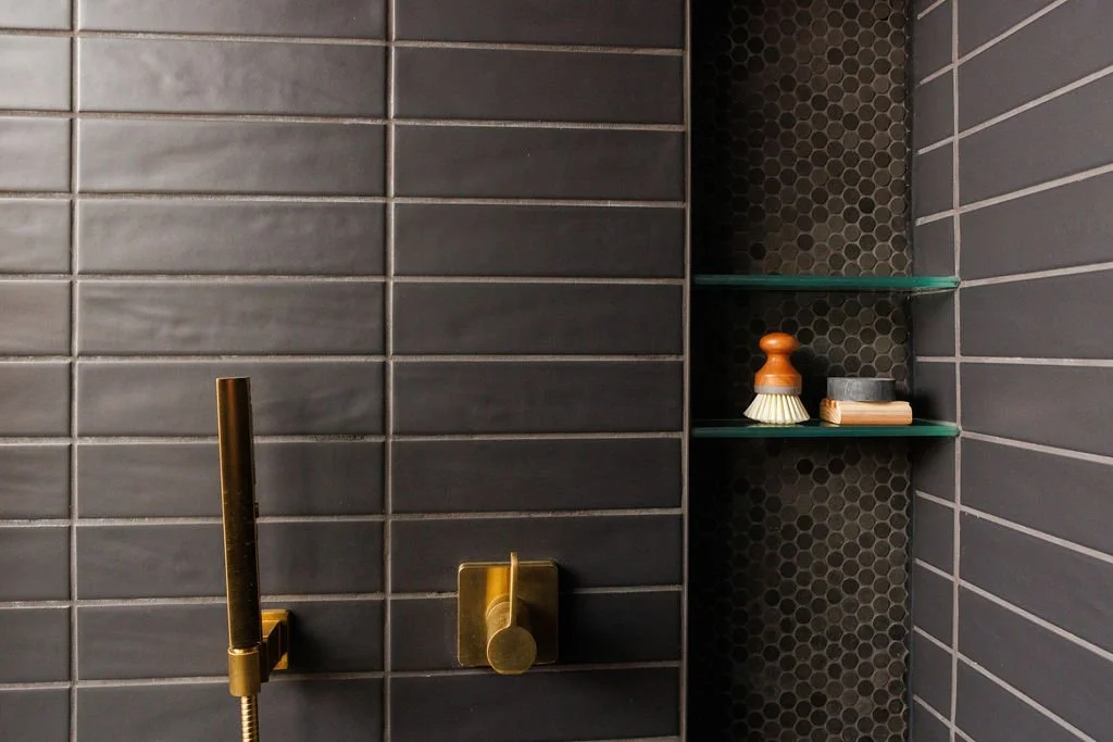 Close-up of a modern bathroom shower with dark gray subway tiles and two glass shelves holding grooming items, with a brass shower fixture.