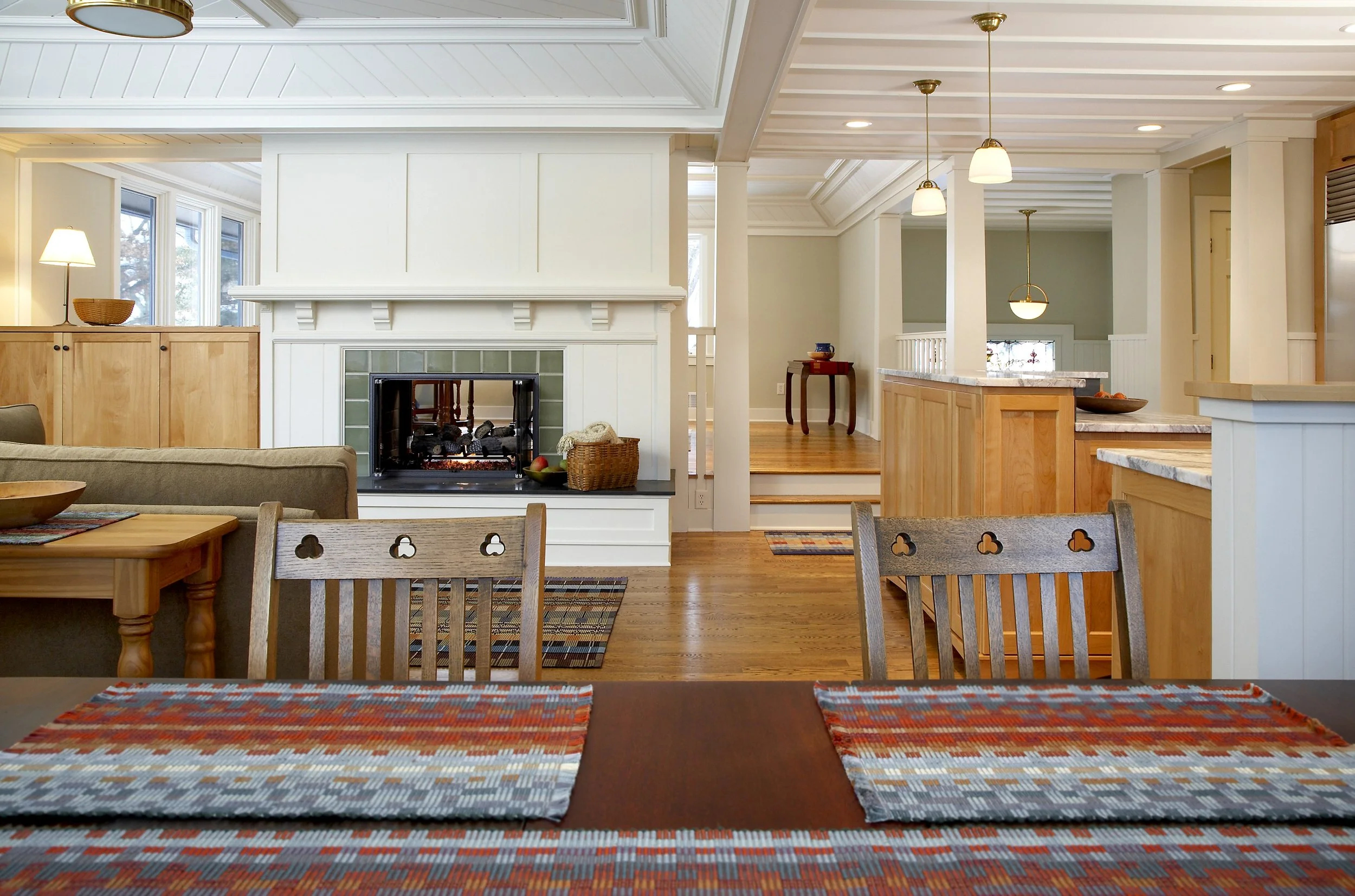 Open-concept living room with fireplace, wooden floors, and kitchen area visible in the background, decorated in neutral tones with multiple pendant lights.