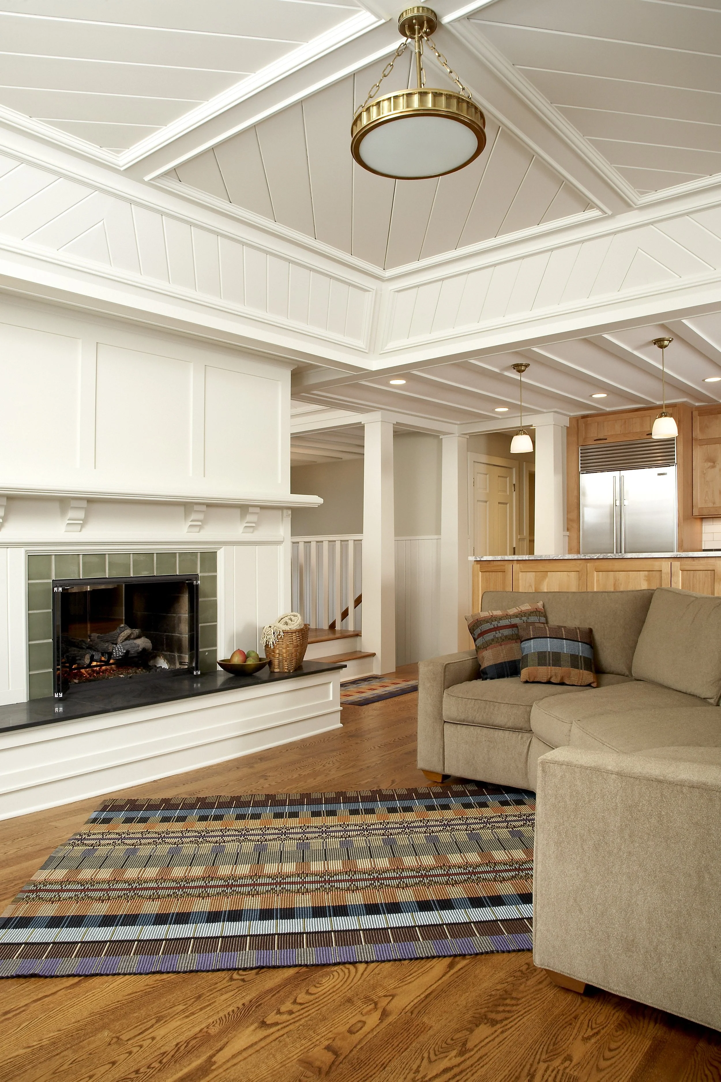Living room with beige sofa, plaid pillows, hardwood floors, colorful striped rug, white fireplace, and kitchen area with wooden cabinets, stainless steel refrigerator, and pendant lights.