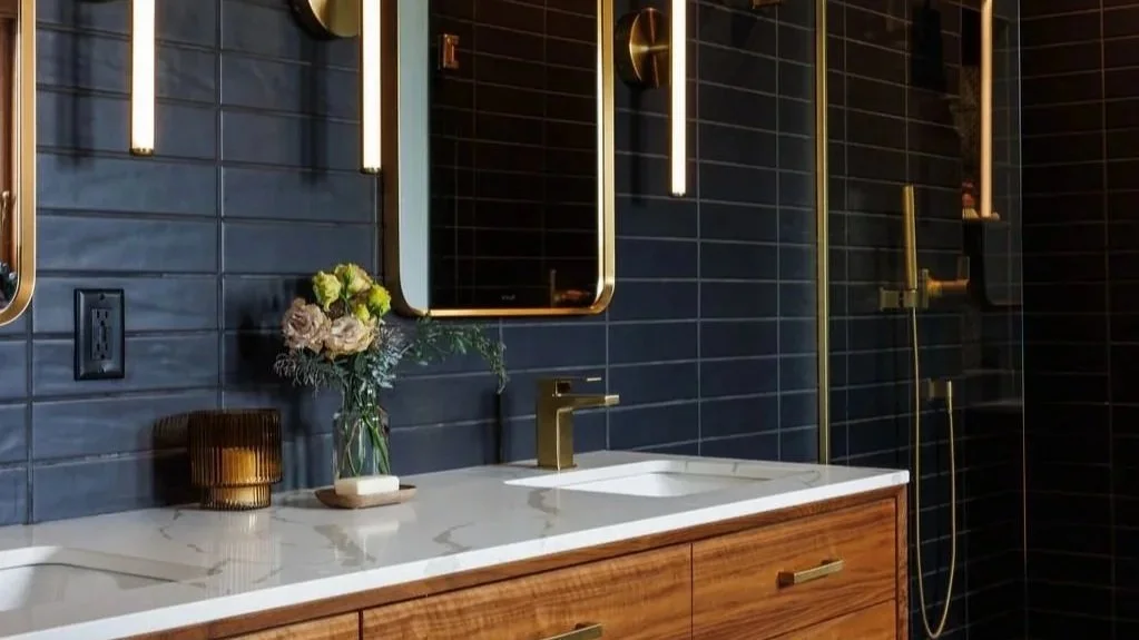 Modern bathroom with dark tiled walls and a wooden vanity with a white marble countertop. Gold fixtures include a faucet, drawer handles, and wall-mounted lights beside mirrors. There is a vase with flowers and a glass candle holder on the vanity. A 