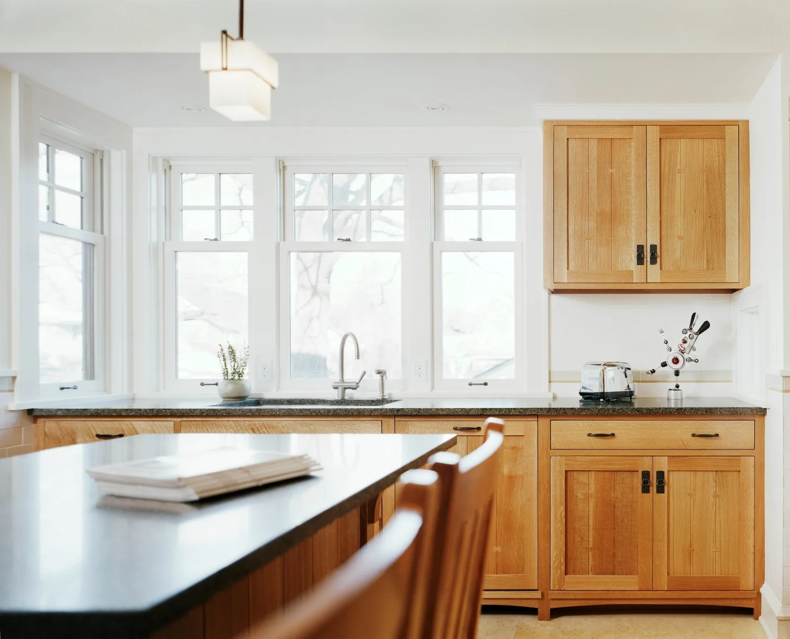 Kitchen with wooden cabinets, granite countertop, white backsplash, four large windows, a small potted plant, a kitchen sink, and appliances including a toaster and a utensil holder shaped like a robot hand.