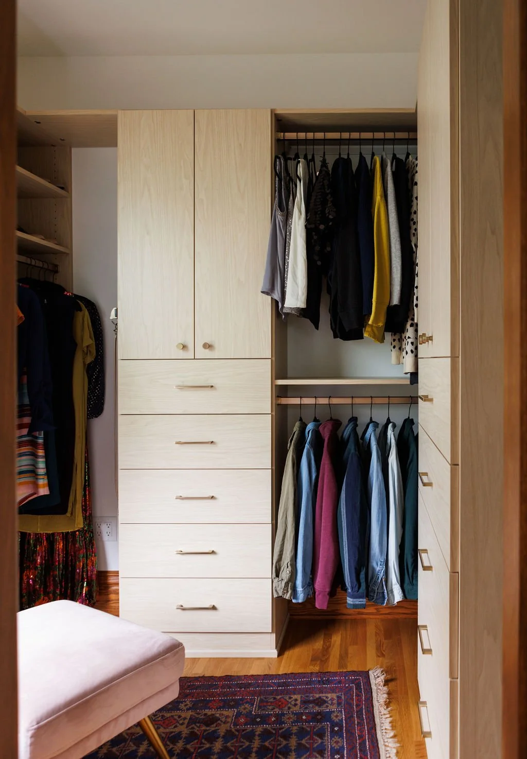 A walk-in closet with built-in wardrobes and hanging clothes, a pink cushioned stool, and a patterned rug on a hardwood floor.