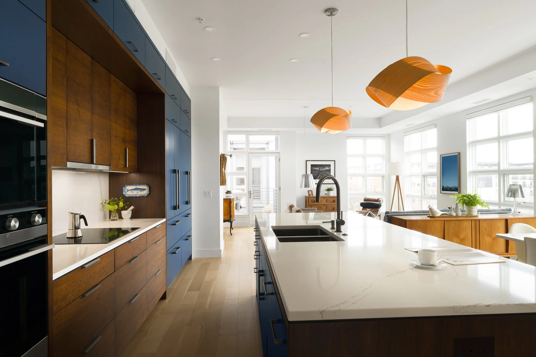 Modern open-concept kitchen with wooden and blue cabinetry, white marble island, large windows letting in natural light, and contemporary lighting fixtures.
