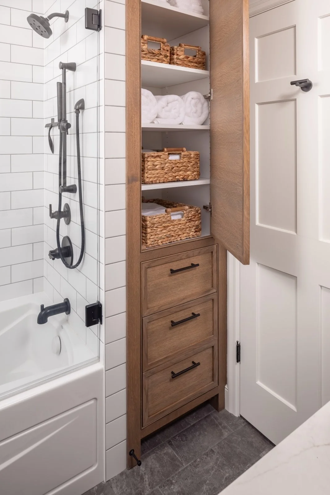 Bathroom with a white tiled shower area and a wooden cabinet with open shelves containing rolled towels and woven baskets.