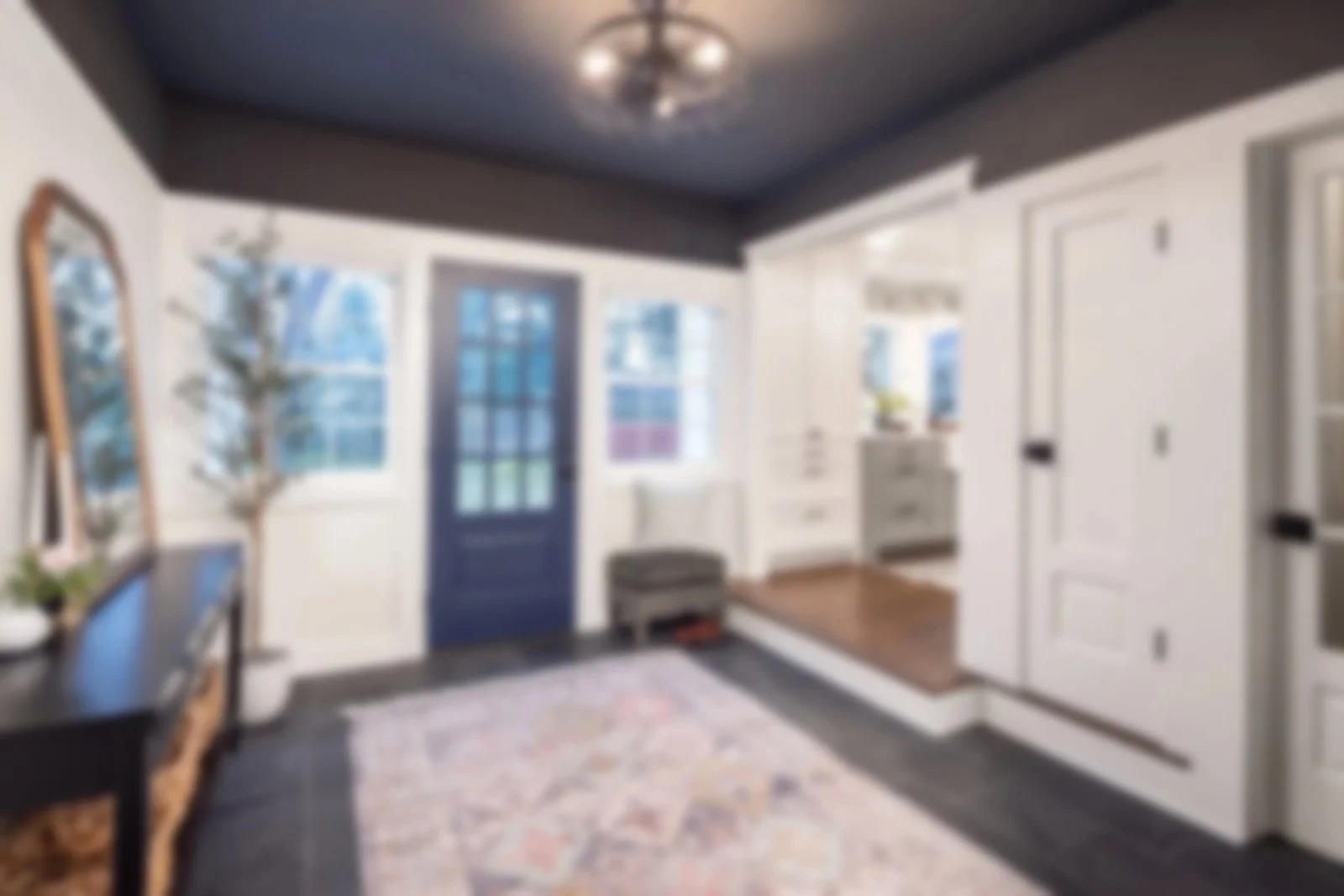 A blurred image of a cozy, well-lit entryway or mudroom with a dark ceiling, white walls, and hardwood floor. There's a blue door, a rug, and various furniture and decorative items, including a mirror, a small table, and a white chest of drawers.