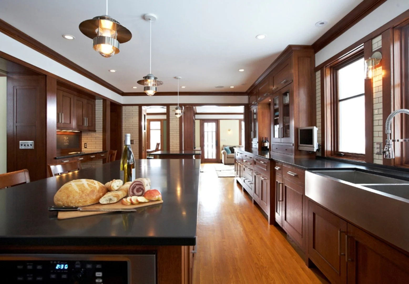 Modern kitchen with dark wood cabinets, black countertop, and a large island with bread and wine on a cutting board. Large windows, hardwood floors, and a view into a living area with a sofa and front door in the distance.