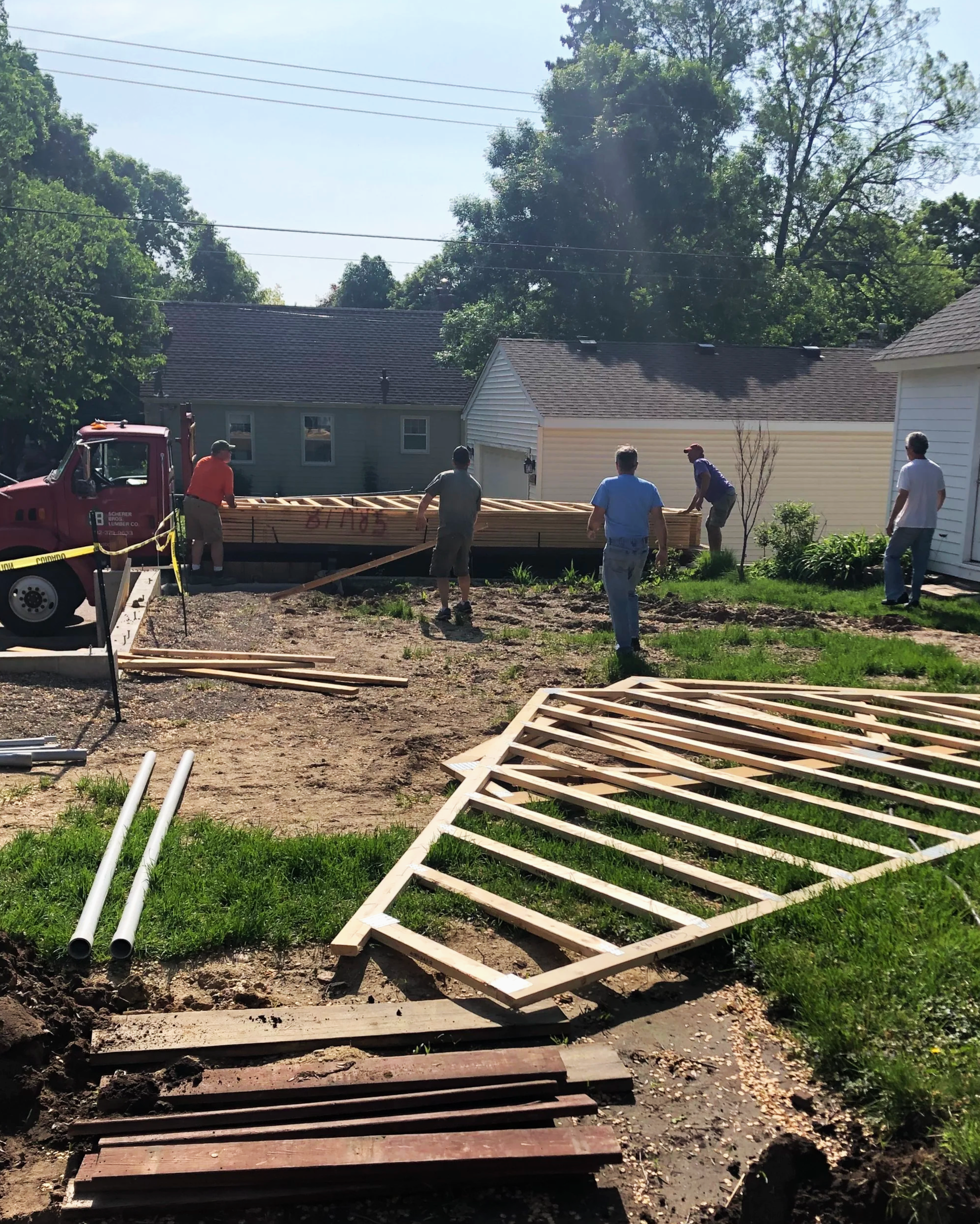 People working on building a wooden deck or porch outside in a residential backyard, with house and trees in the background.