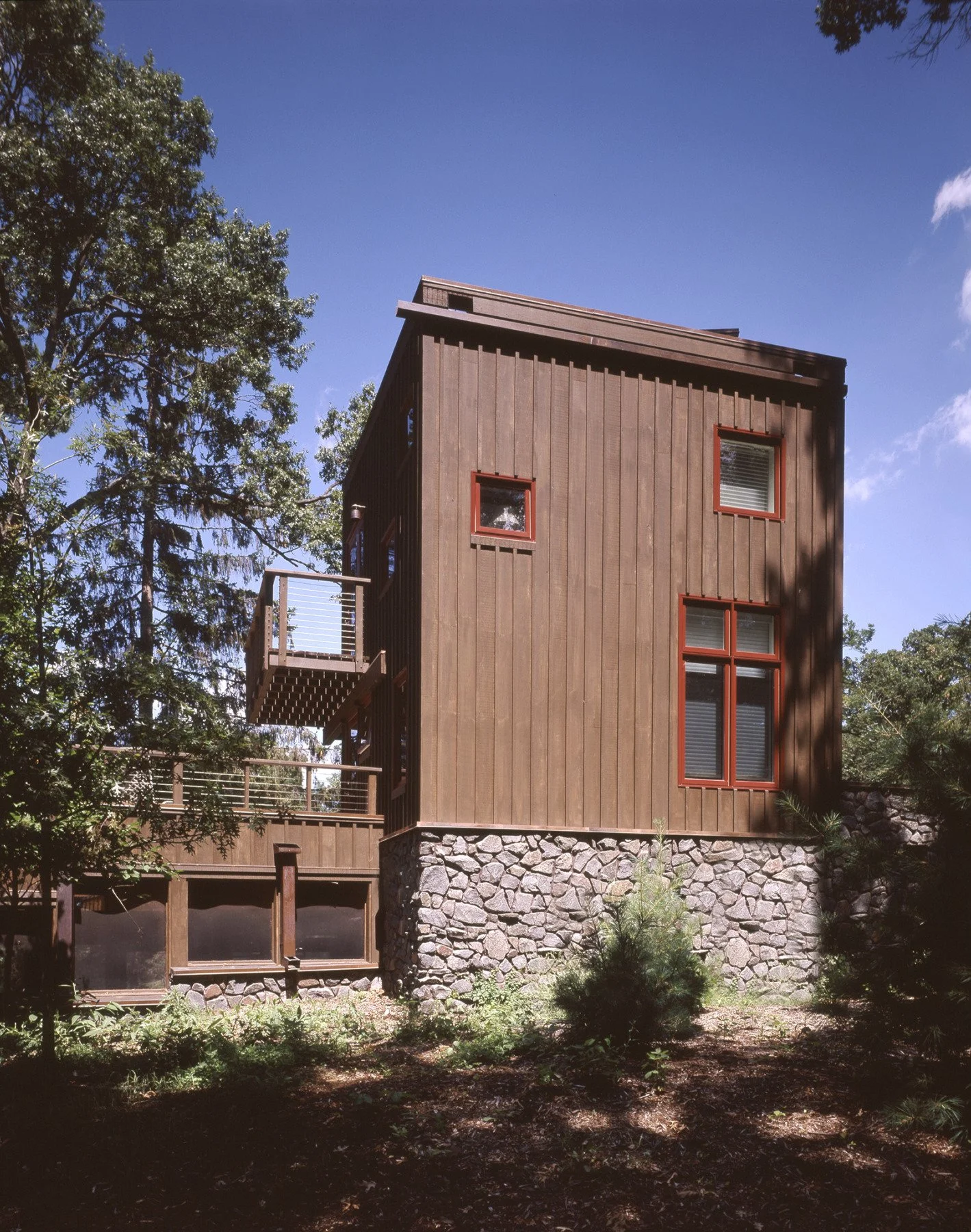 A modern two-story house with a wooden exterior, red window frames, and a stone foundation, surrounded by tall trees and a clear blue sky.