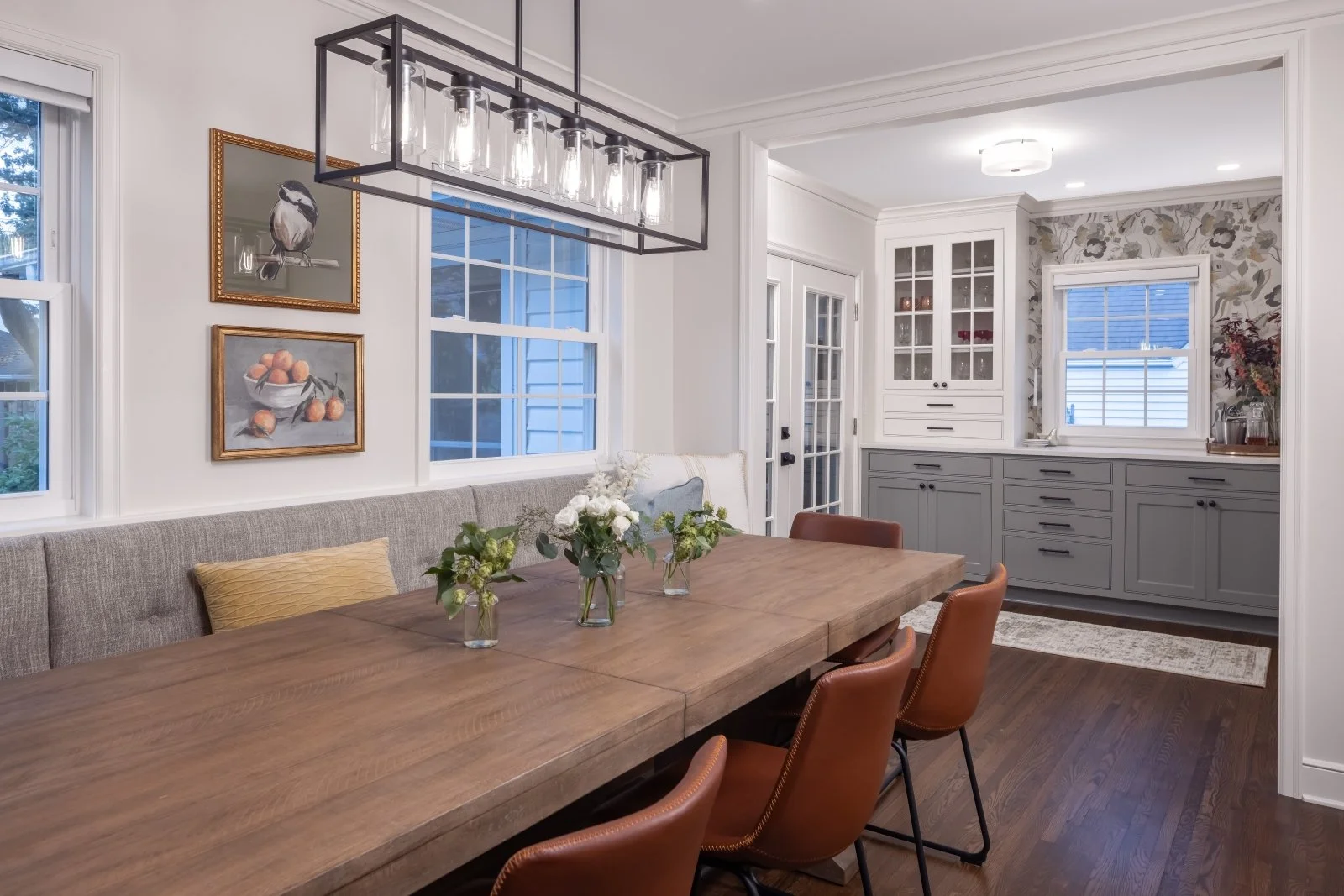 Dining room featuring a wood table with four brown leather chairs, a bench with cushions, floral arrangements, white walls, two windows, wall art, and a kitchen nook with gray cabinets and floral wallpaper.