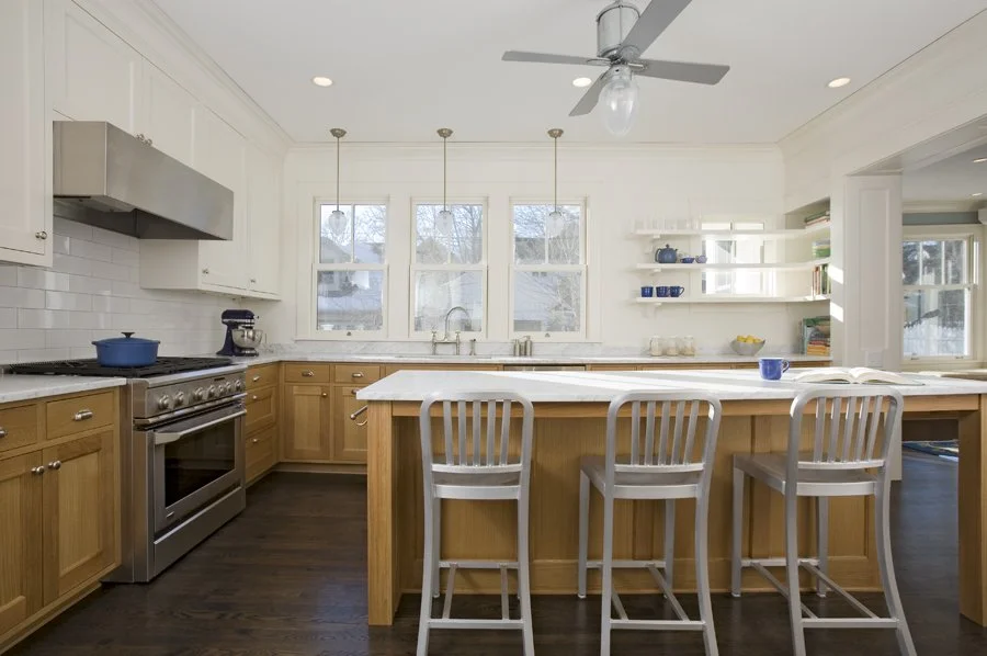 Bright white kitchen with natural wood cabinets, a large island with a white countertop, three white chairs, windows with a view of trees, and open shelving with dishes and books.