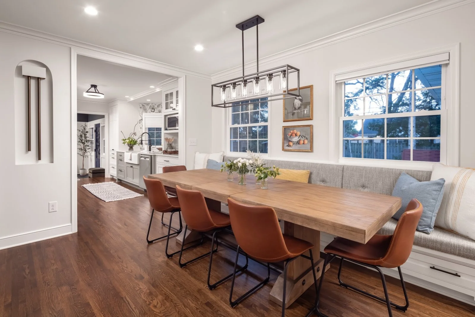 A dining room with a wooden table, leather chairs, and a gray cushioned bench with pillows, overlooking a kitchen with white cabinets and window views of trees outside.