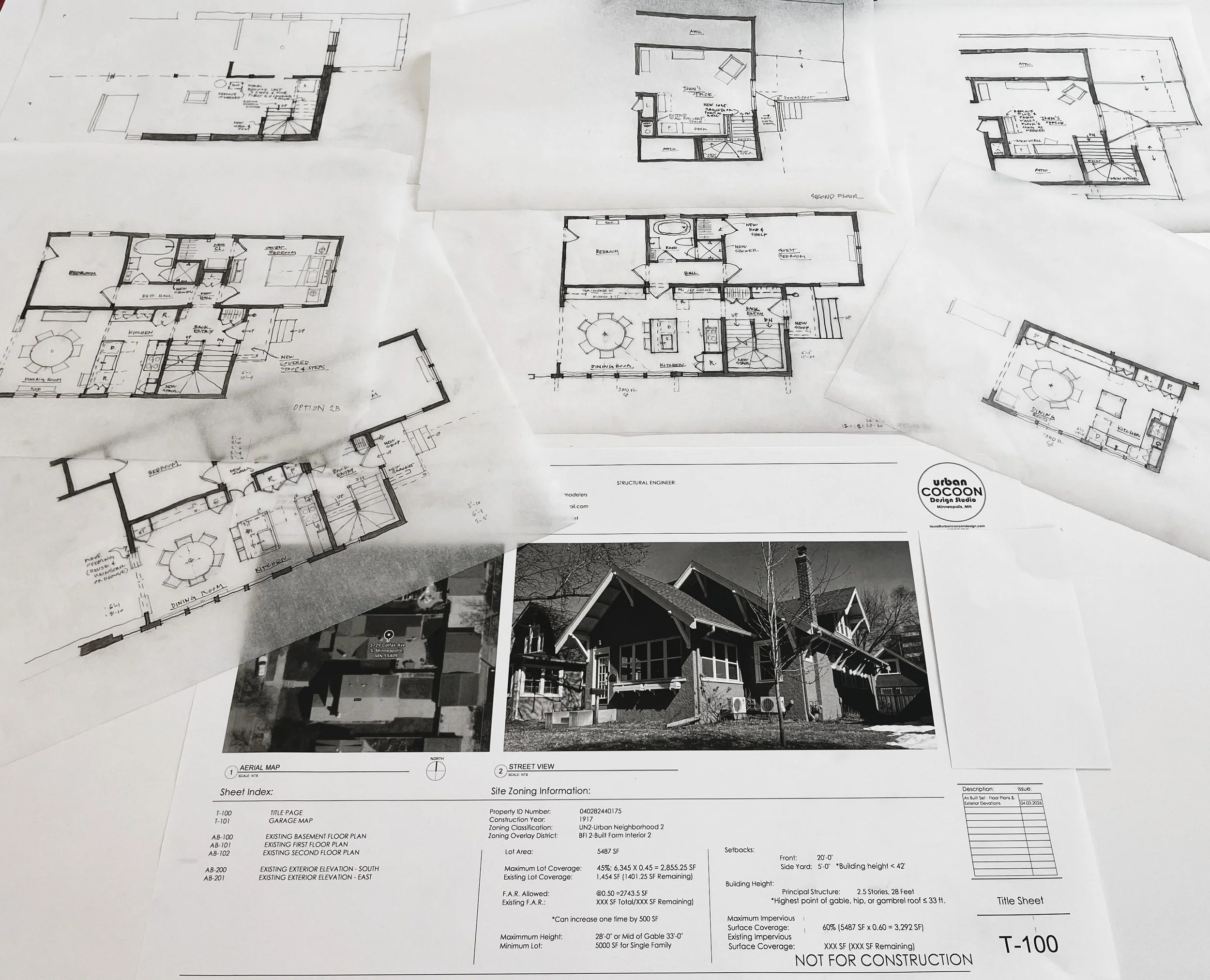 A collection of architectural blueprints and house plans spread out on a table, with some black-and-white photos of houses in the background.