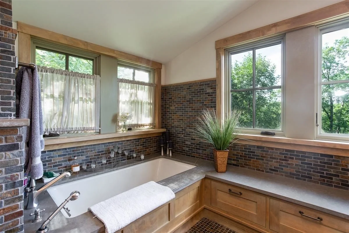 A bathroom with a large soaking tub next to two windows, brick walls, and wooden accents, with a potted plant on the counter.