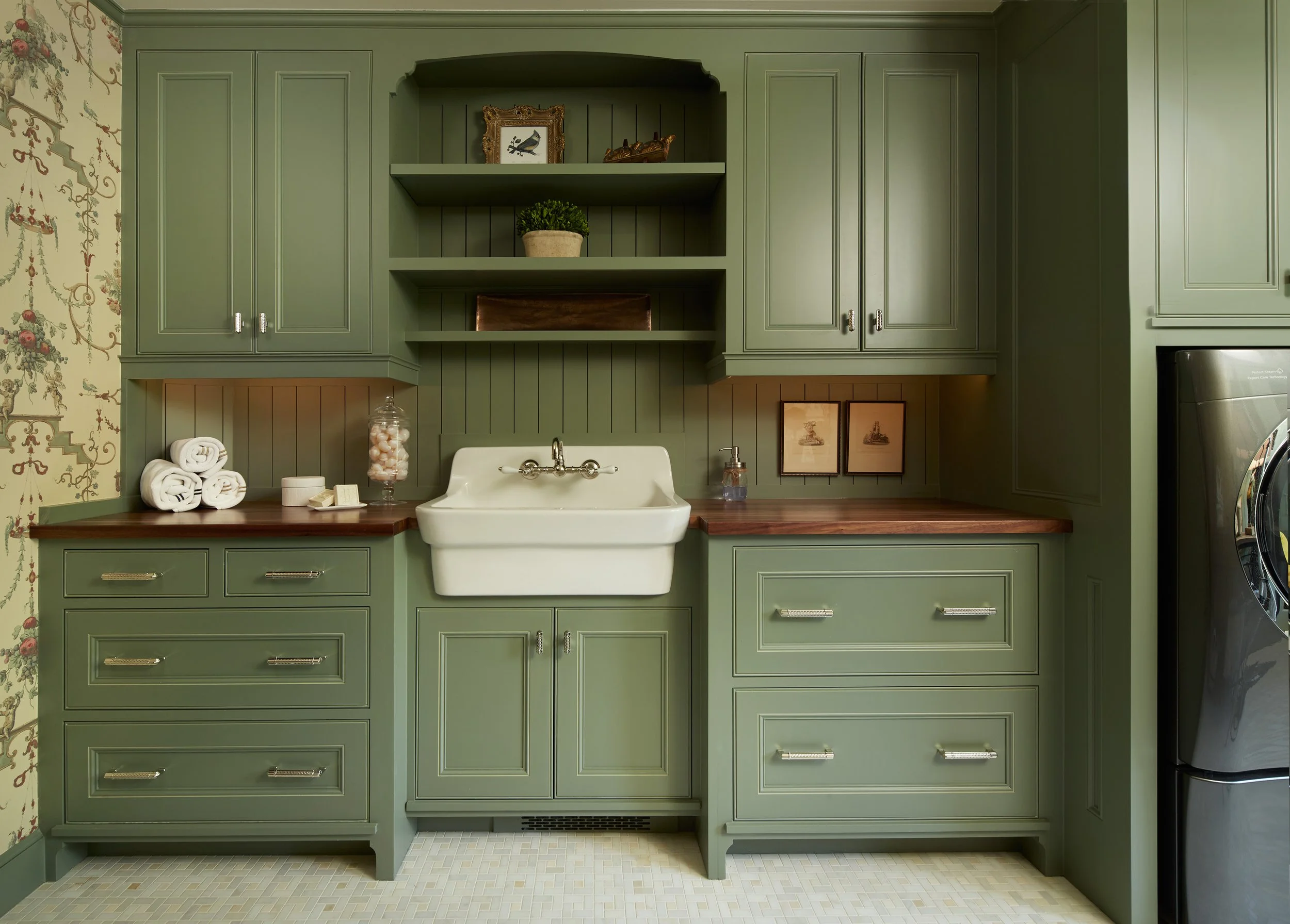 Green kitchen cabinets with open shelves, a farmhouse sink, rolled white towels, framed pictures, and decorative items on the countertop.