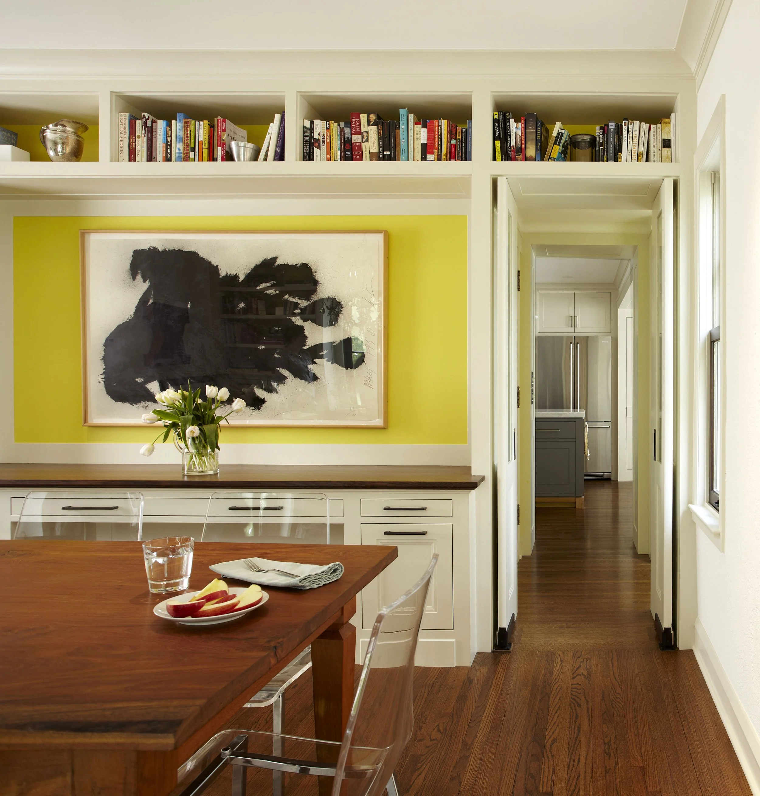 A kitchen and dining area with a wooden table set with a glass of water, a napkin, and sliced apples. Behind the table is a white cabinet with drawers, above which is a bright yellow wall decorated with black abstract artwork. The space extends into 
