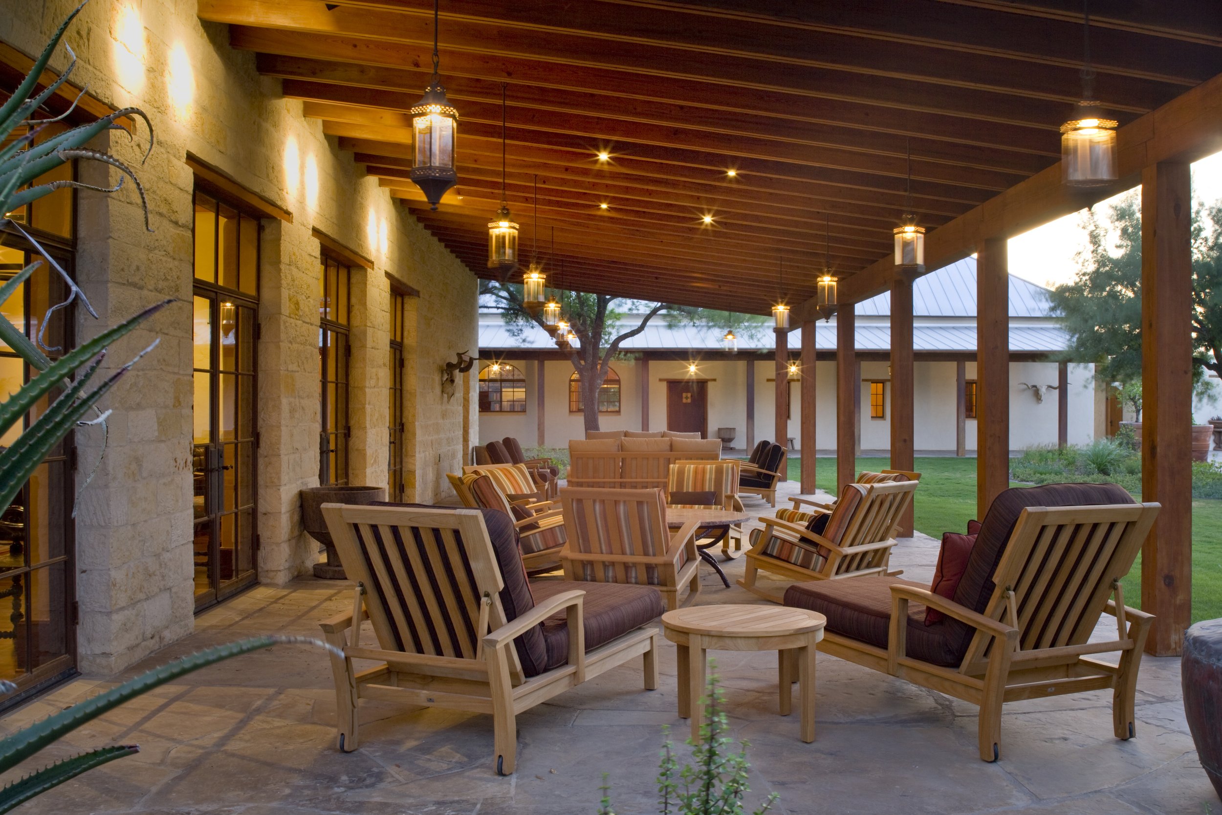 Covered outdoor patio with wooden ceiling, hanging lanterns, and outdoor furniture including chairs and tables, overlooking a green yard with trees.