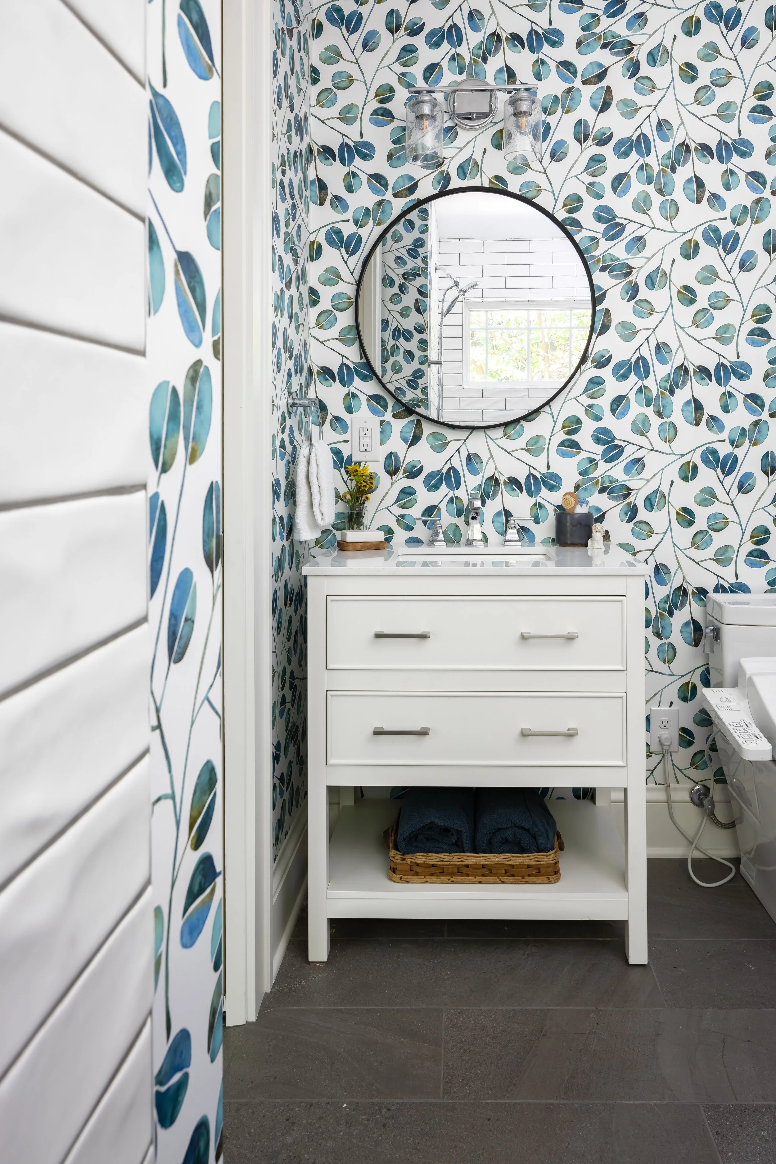A bathroom with wallpaper featuring blue and green leaves, a white vanity with a round mirror above, and a window reflected in the mirror.