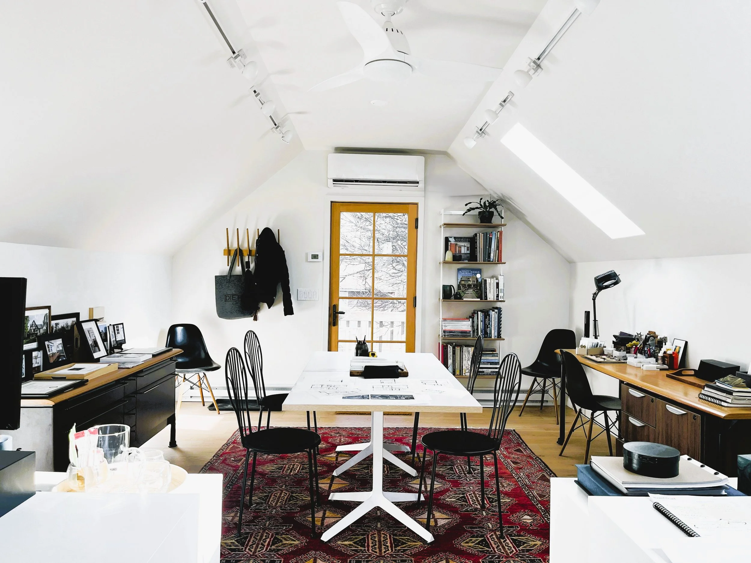 A cozy home office with white walls and sloped ceiling, featuring a large window with snowy landscape outside, a central white table surrounded by black chairs, bookshelves with plants and books, and various office supplies, all situated on a patterned red rug.