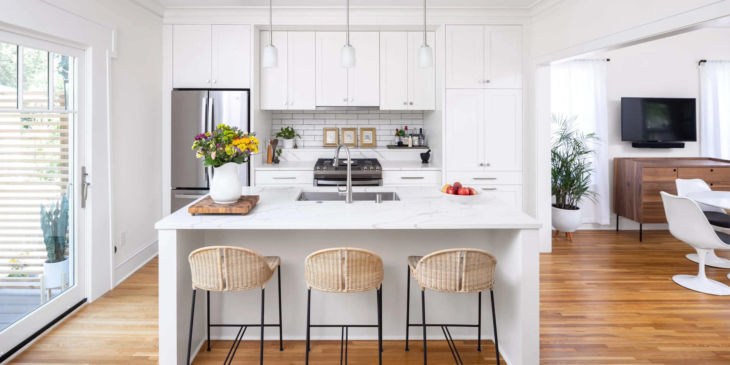 A modern white kitchen with a marble island, pendant lights, a fridge, a stove, and decor. To the right, there's a living area with a wall-mounted TV, a wooden cabinet, a large potted plant, and a dining table with white chairs.