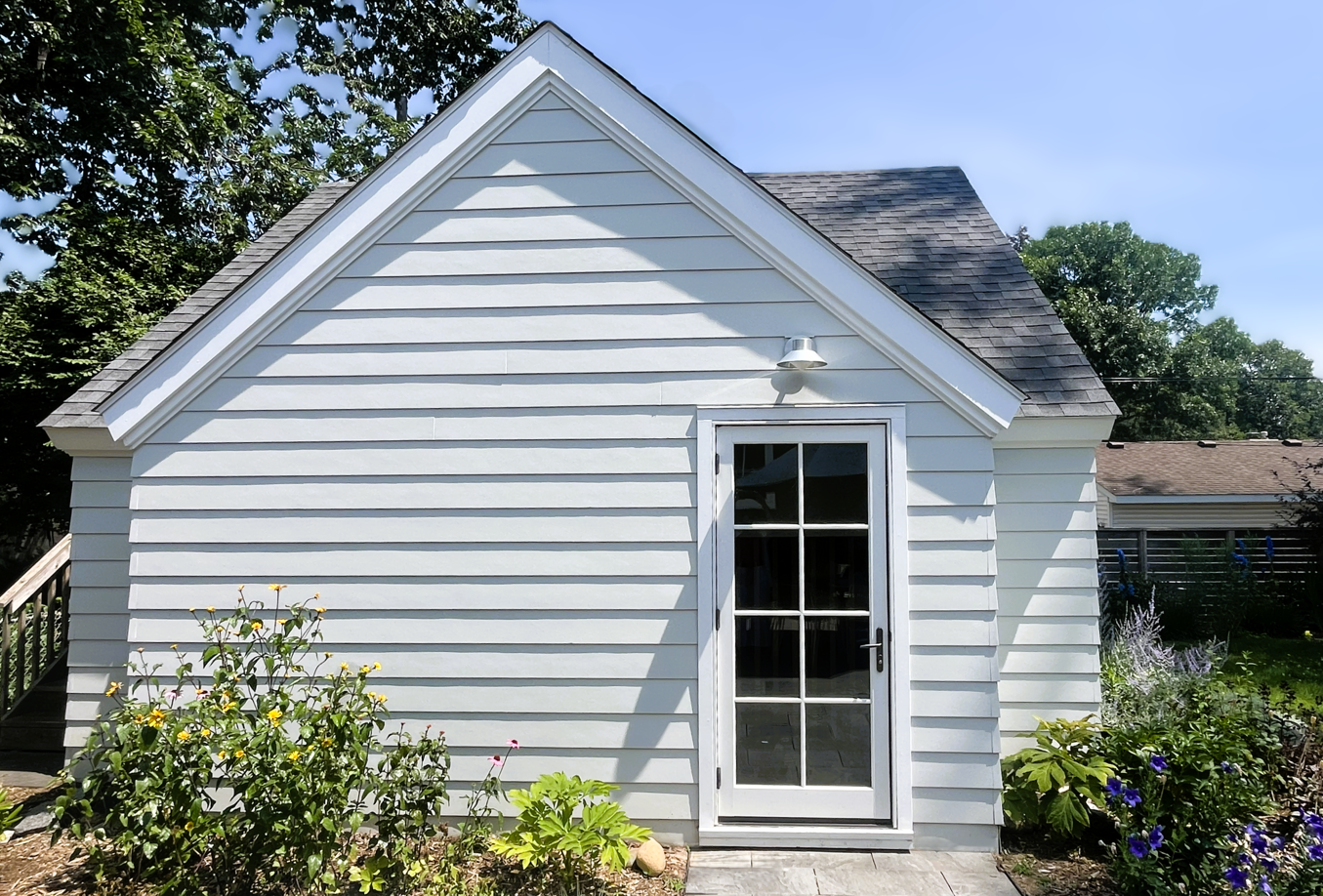 Small white house with a gabled roof, exterior light fixture, and a glass-paneled door, surrounded by flowers and bushes under a clear blue sky.