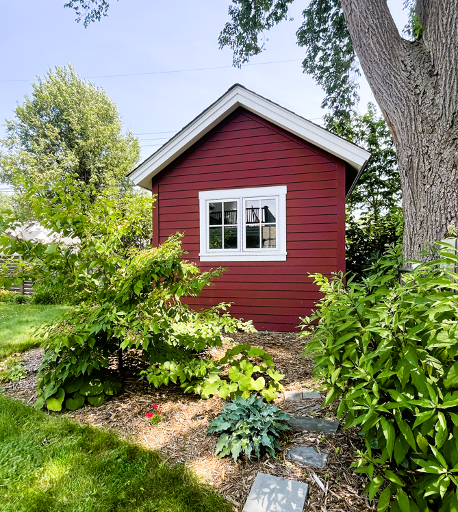 Small red shed with white window in a lush garden with green bushes, a large tree, and stone pathway.
