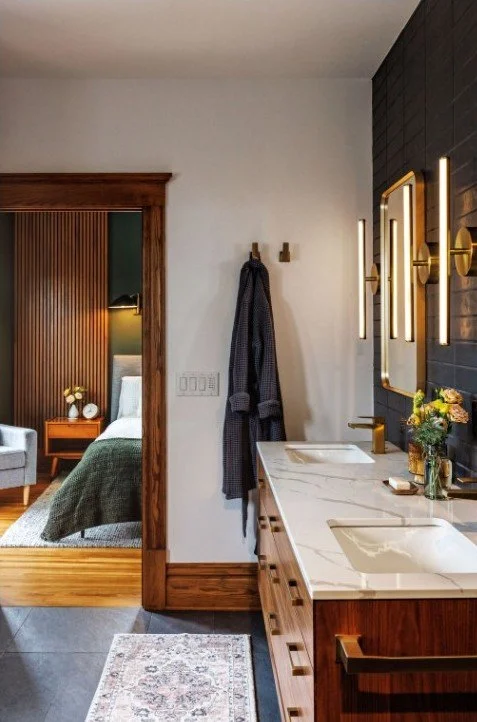 Modern bathroom with white walls, a marble countertop, two sinks, and brass fixtures, adjacent to a bedroom visible through a doorway.